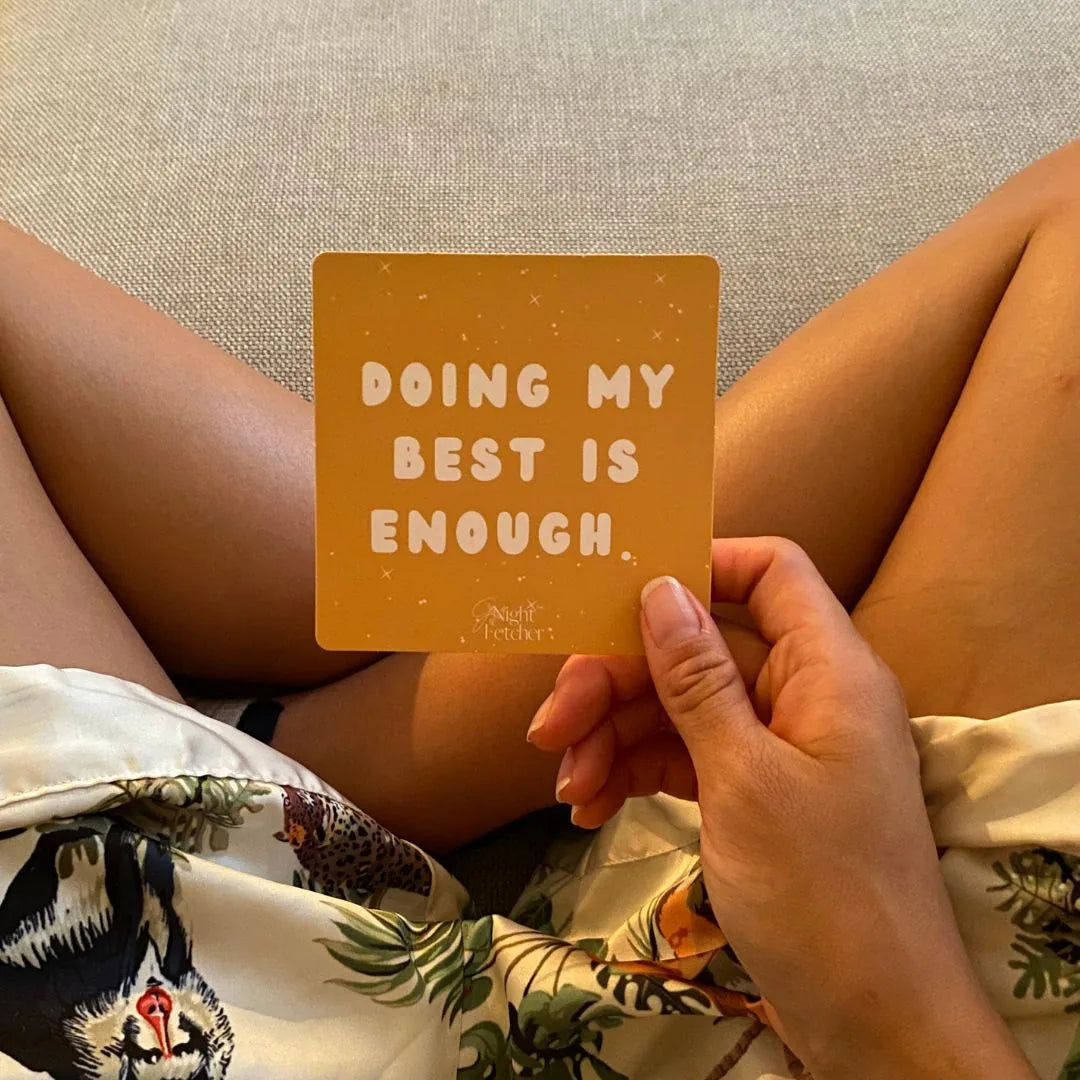 Yellow affirmation card reading "DOING MY BEST IS ENOUGH" held in a hand, with patterned fabric and legs visible.