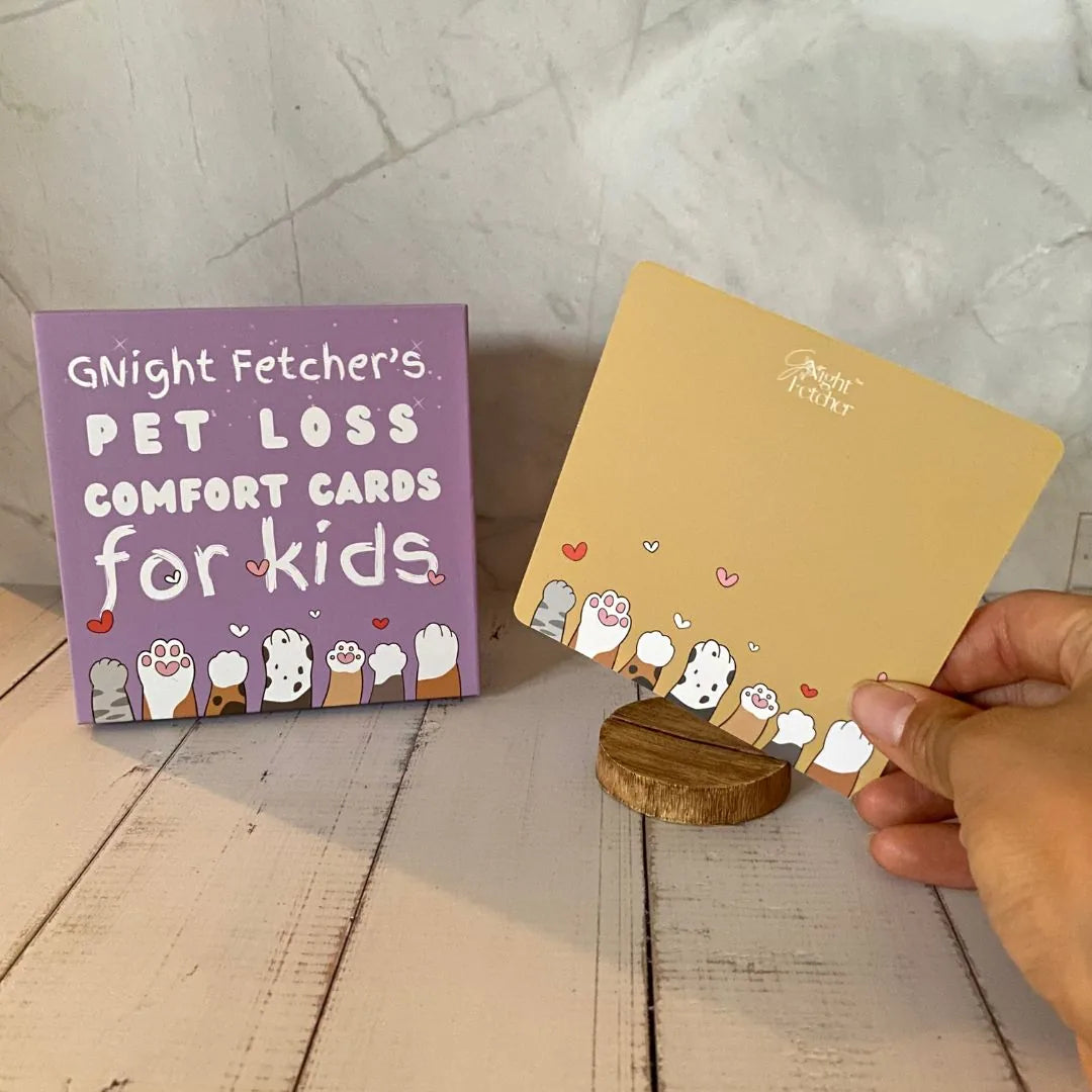 Purple box of "Pet Loss Comfort Cards for Kids" with playful graphics, next to a beige card held by a hand.