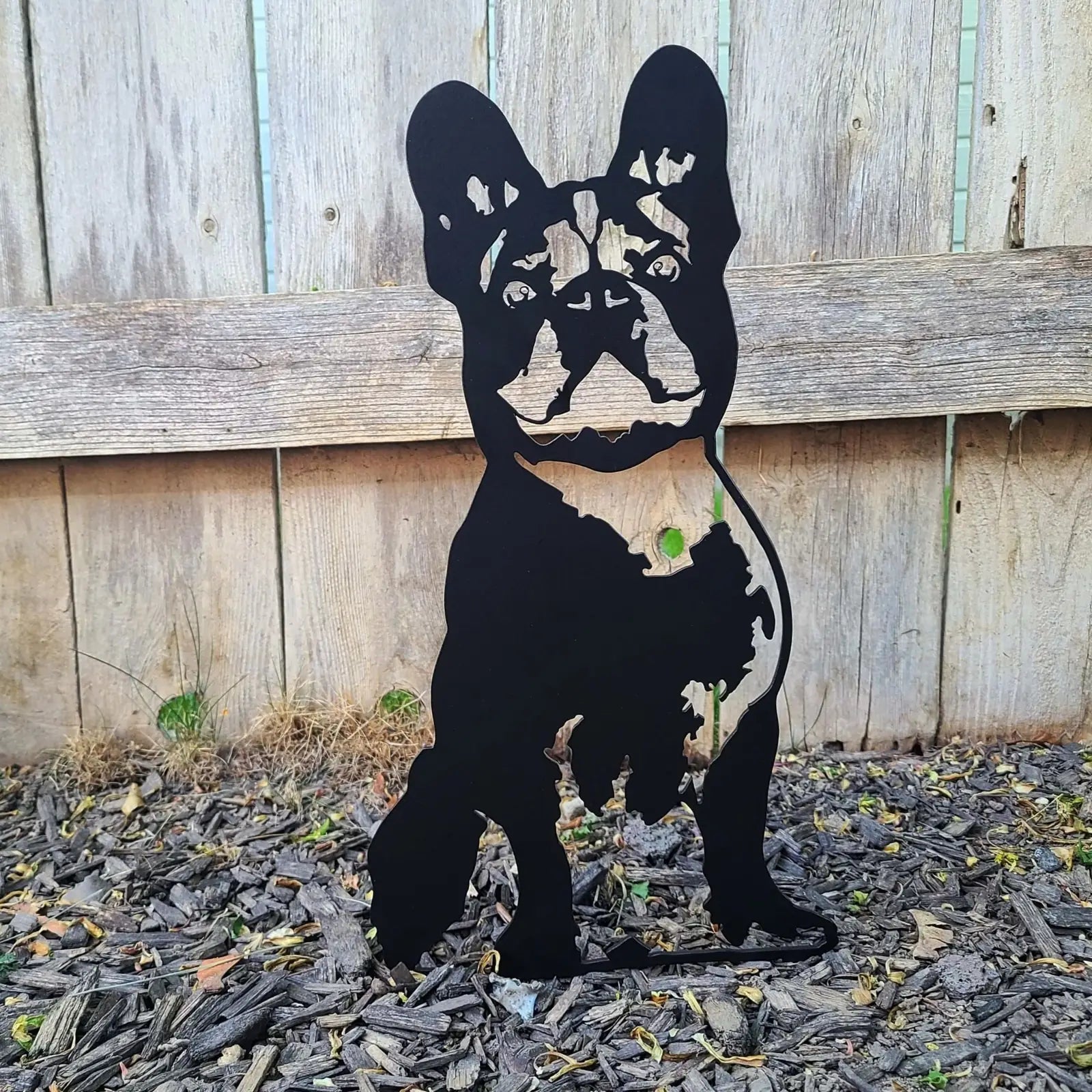 Handmade black metal art silhouette of a French Bulldog standing against a wooden fence, perfect for dog lovers.