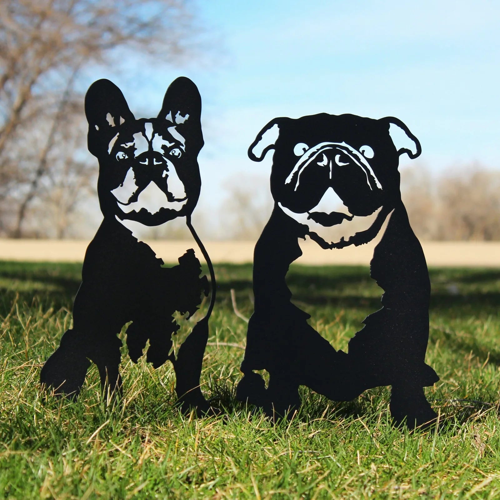 Black metal silhouettes of a French Bulldog and a pug, standing on green grass with a clear sky backdrop.