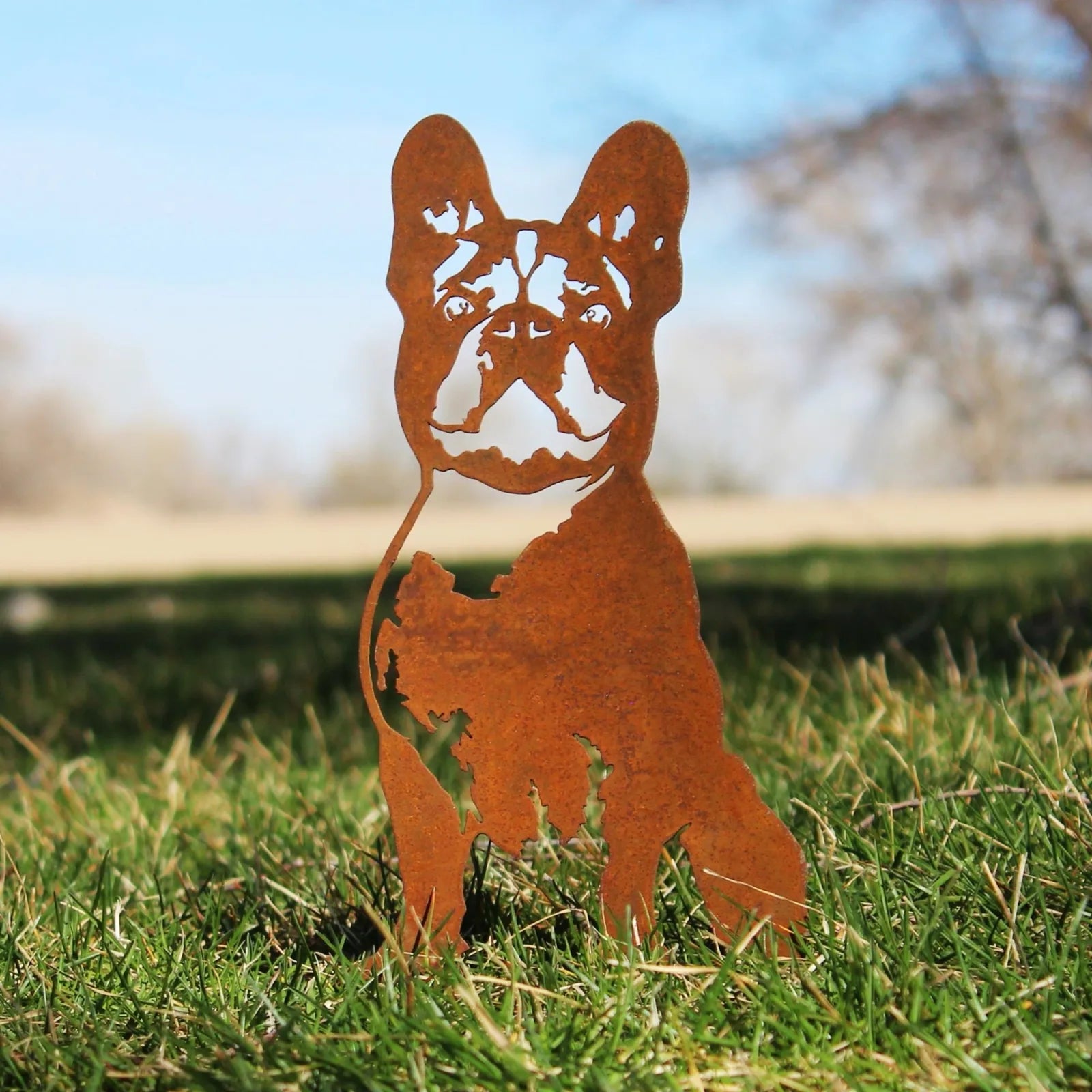 Rust-colored French Bulldog silhouette made of weathering steel, set against green grass and a clear blue sky.