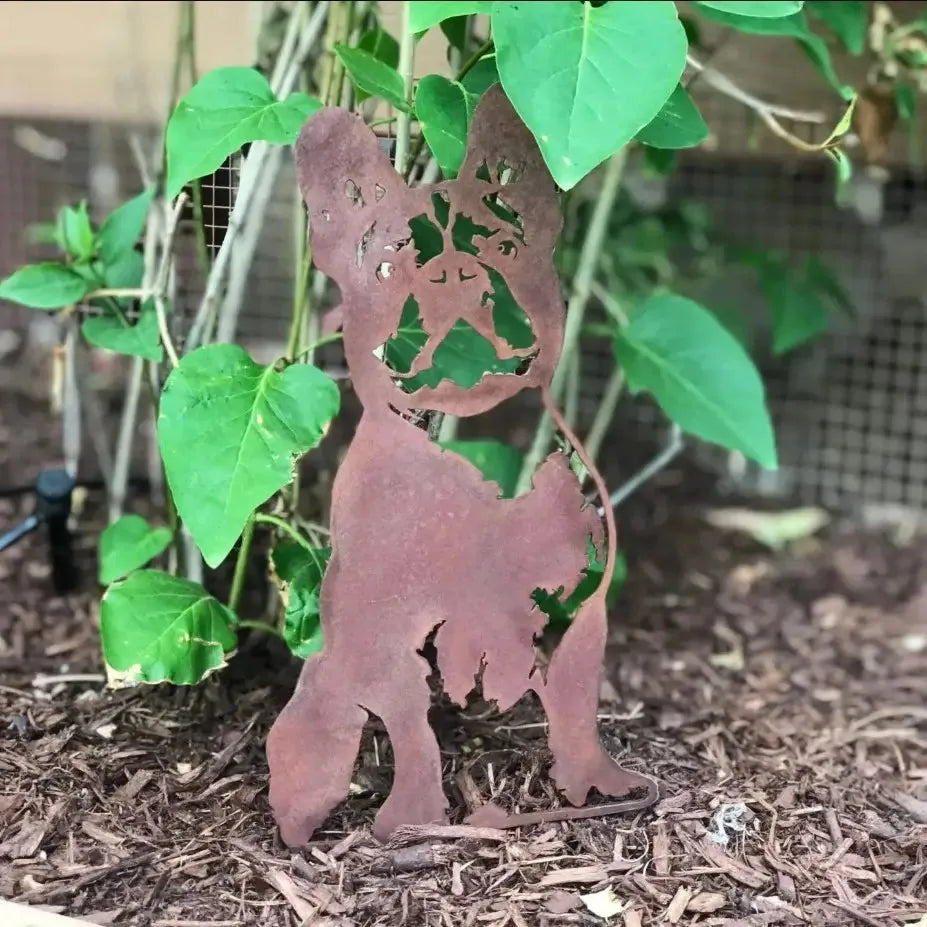 Rusty steel silhouette of a French Bulldog nestled among green leaves, showcasing handmade metal art for dog lovers.
