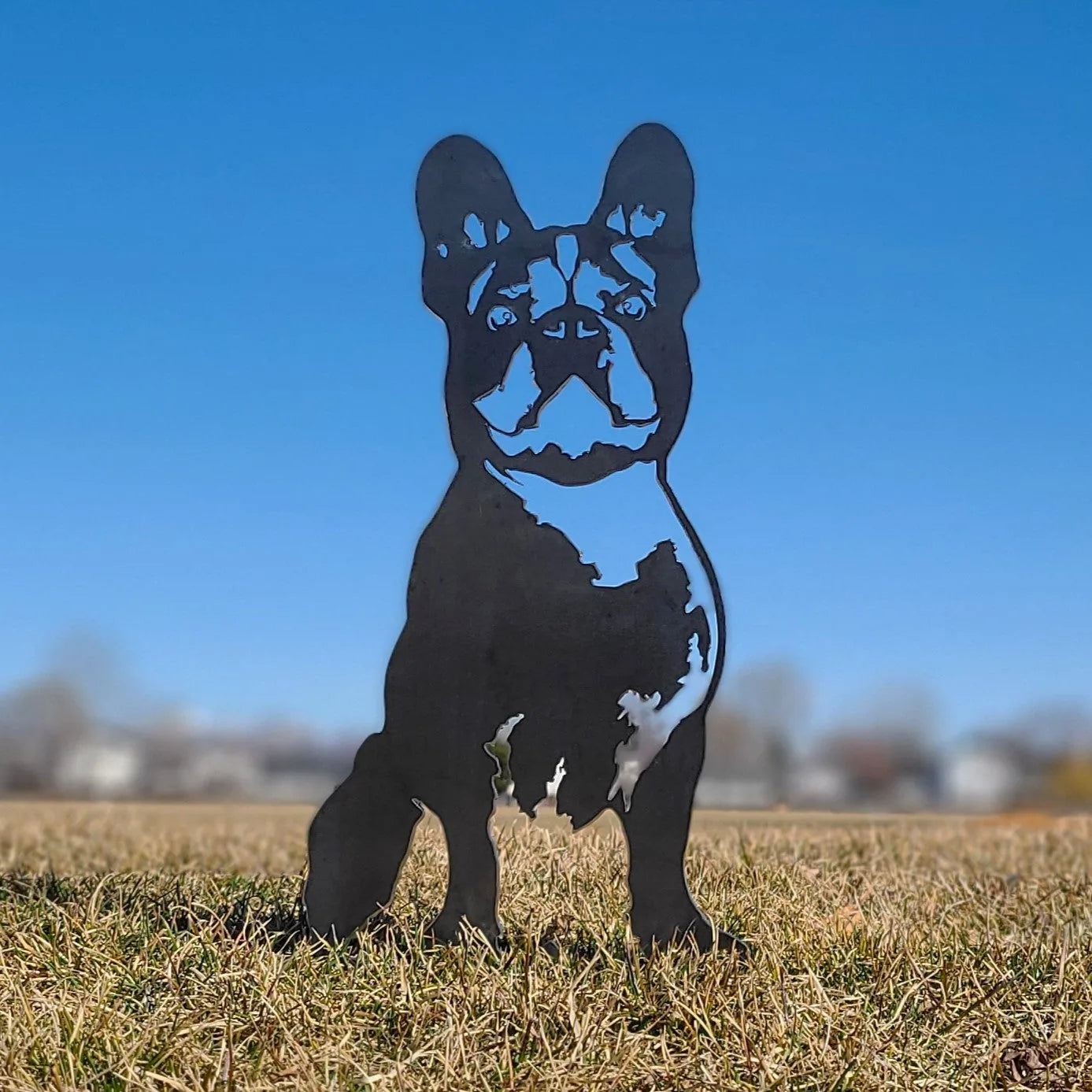 Handmade black metal silhouette of a French Bulldog, set against a clear blue sky and grassy field.