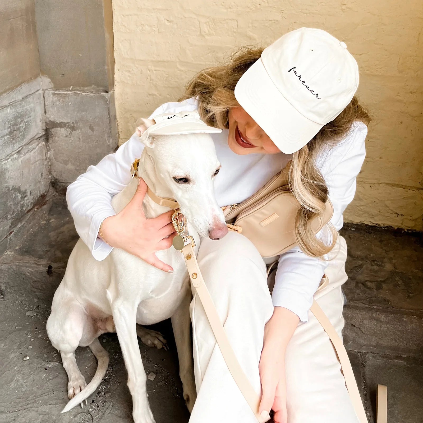 Matching cream baseball hats with "Furever" embroidery, worn by a woman and her white dog, sitting together in a cozy sett...