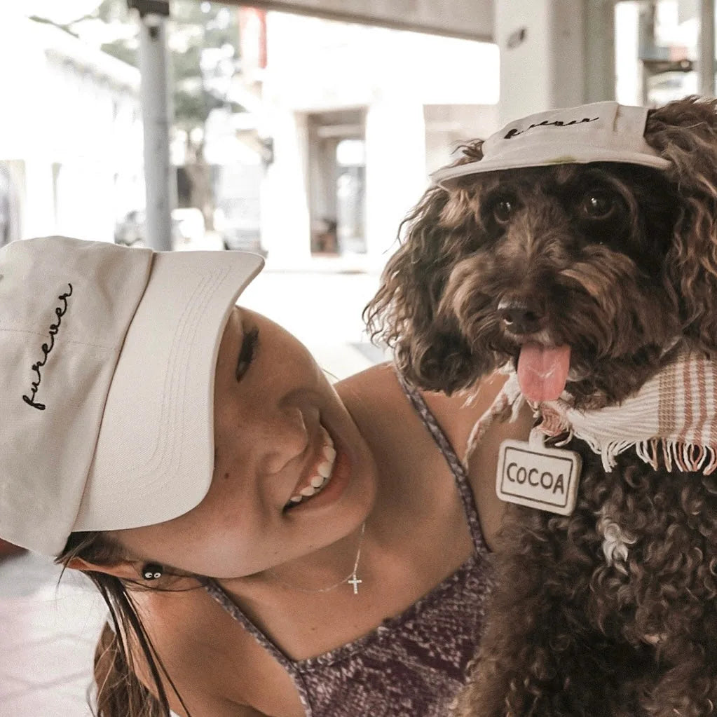 A woman wearing a white baseball cap smiles at her dog, also in a matching white cap, showcasing their "furever" connection.