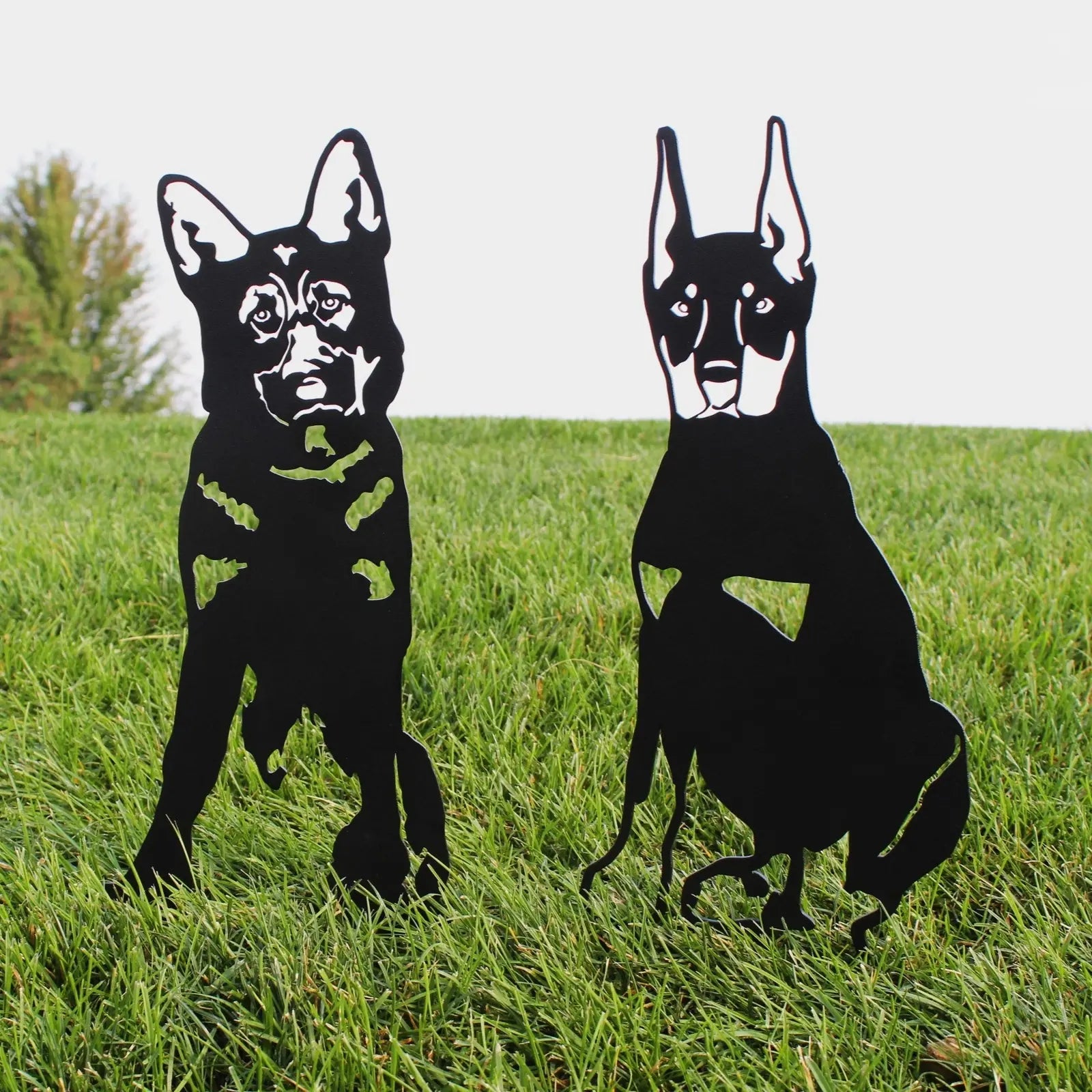 Two black metal dog silhouettes representing a German Shepherd and another breed, set against a grassy background.