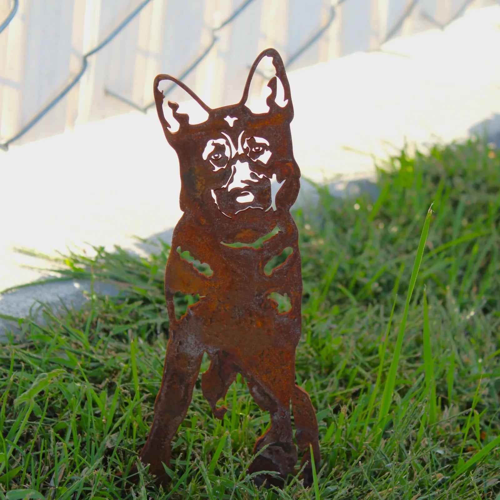 Rust-colored German Shepherd metal silhouette stands in green grass, showcasing detailed cutouts against a sunny backdrop.