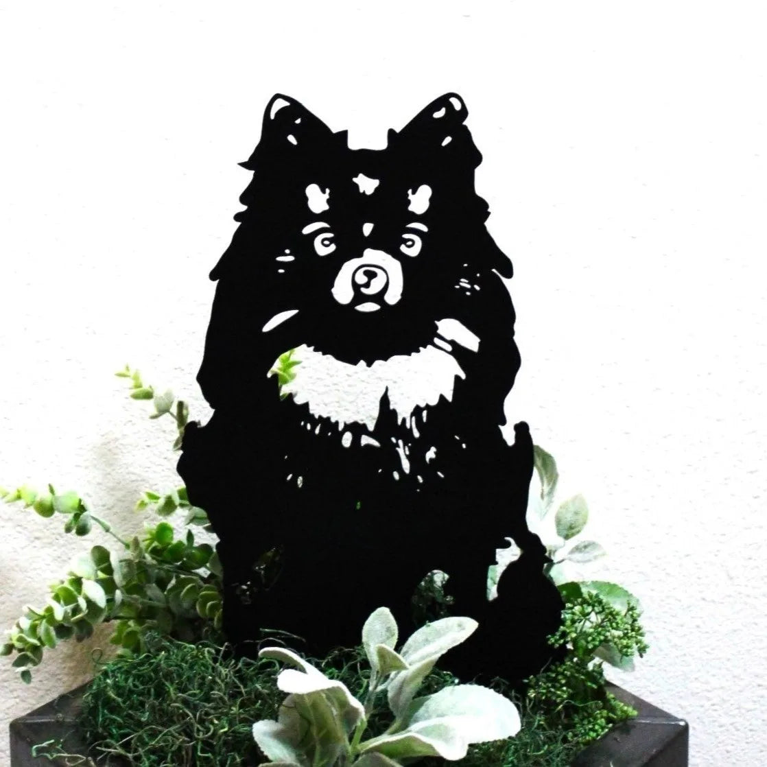Black metal silhouette of a German Spitz dog, surrounded by green foliage for decorative display.