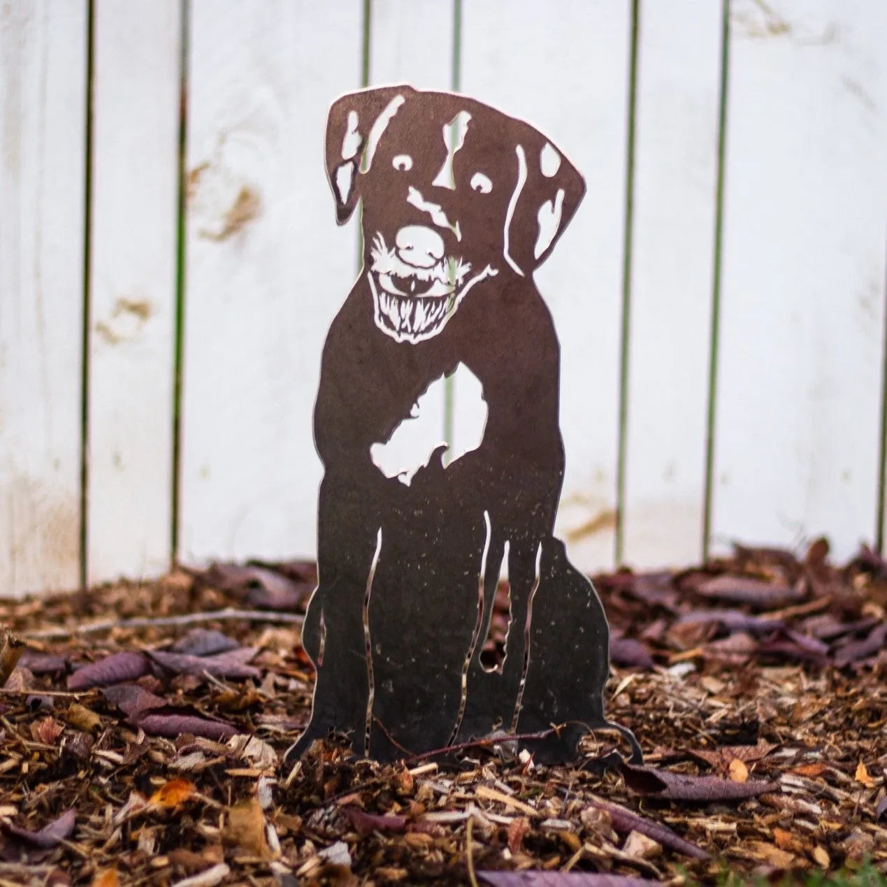 Handmade black metal silhouette of a dog sitting, featured against mulch with a rustic wooden background.