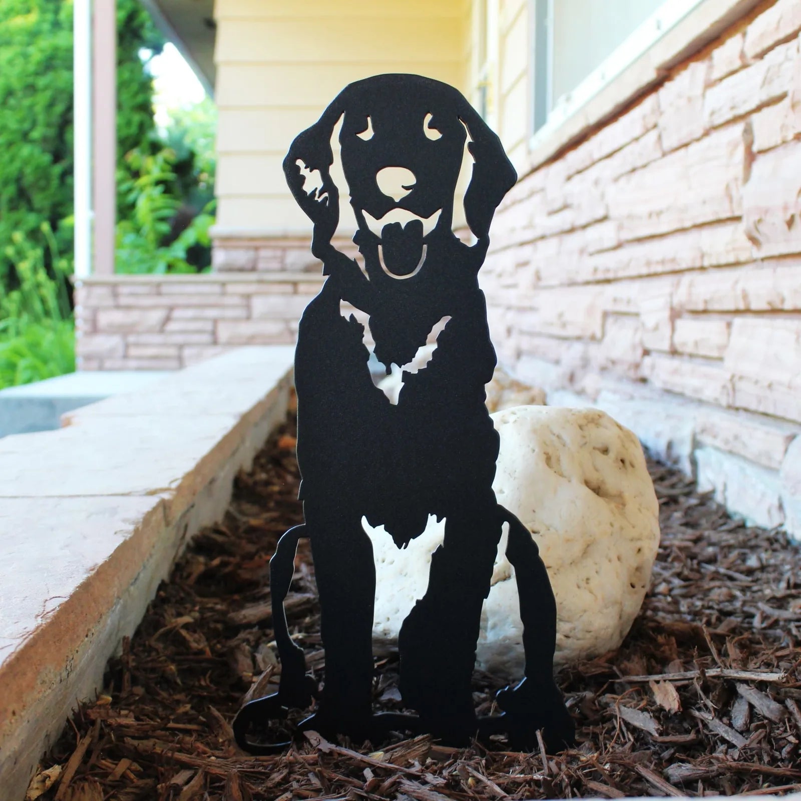 Handcrafted black metal silhouette of a Golden Retriever sitting, showcased in a garden setting with mulch and rocks.