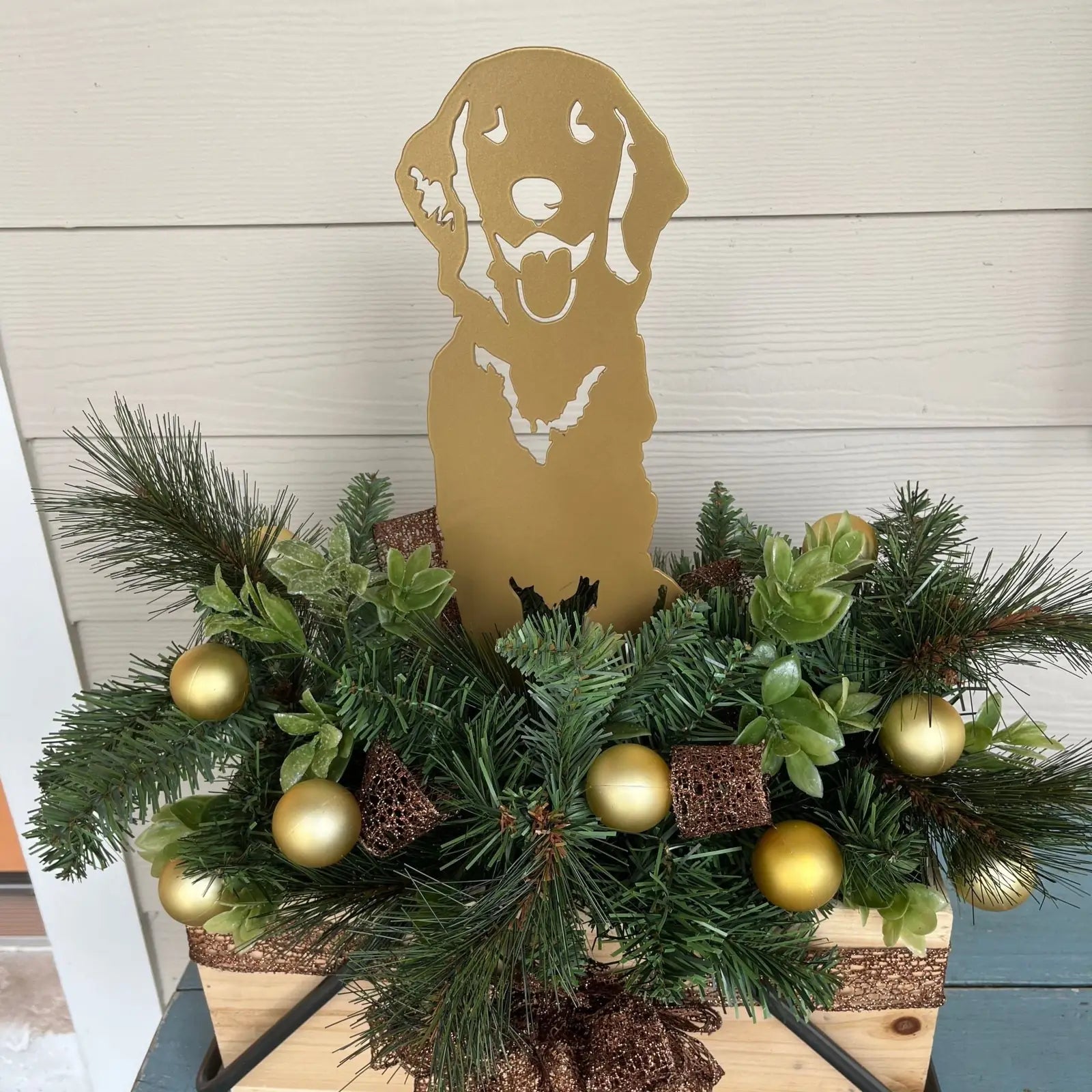 Golden Retriever silhouette in gold metal surrounded by greenery and festive ornaments in a decorative arrangement.