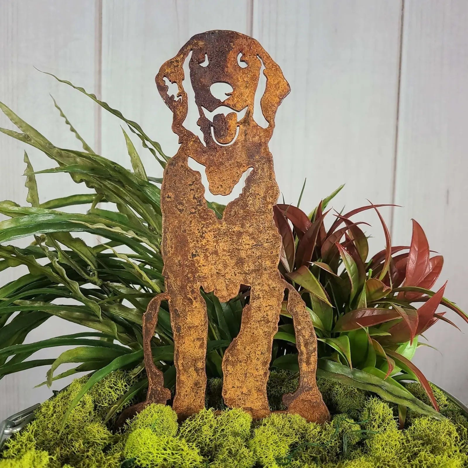 Handcrafted rust-colored metal silhouette of a Golden Retriever sitting amidst vibrant green moss and foliage.