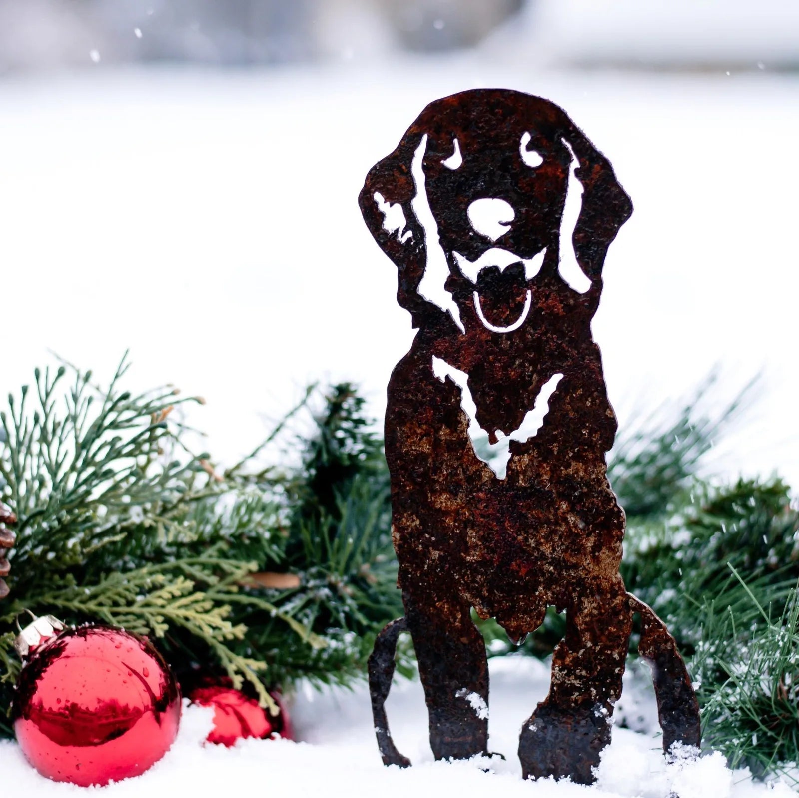 Handcrafted rusted metal silhouette of a Golden Retriever surrounded by green foliage and red ornaments in snow.