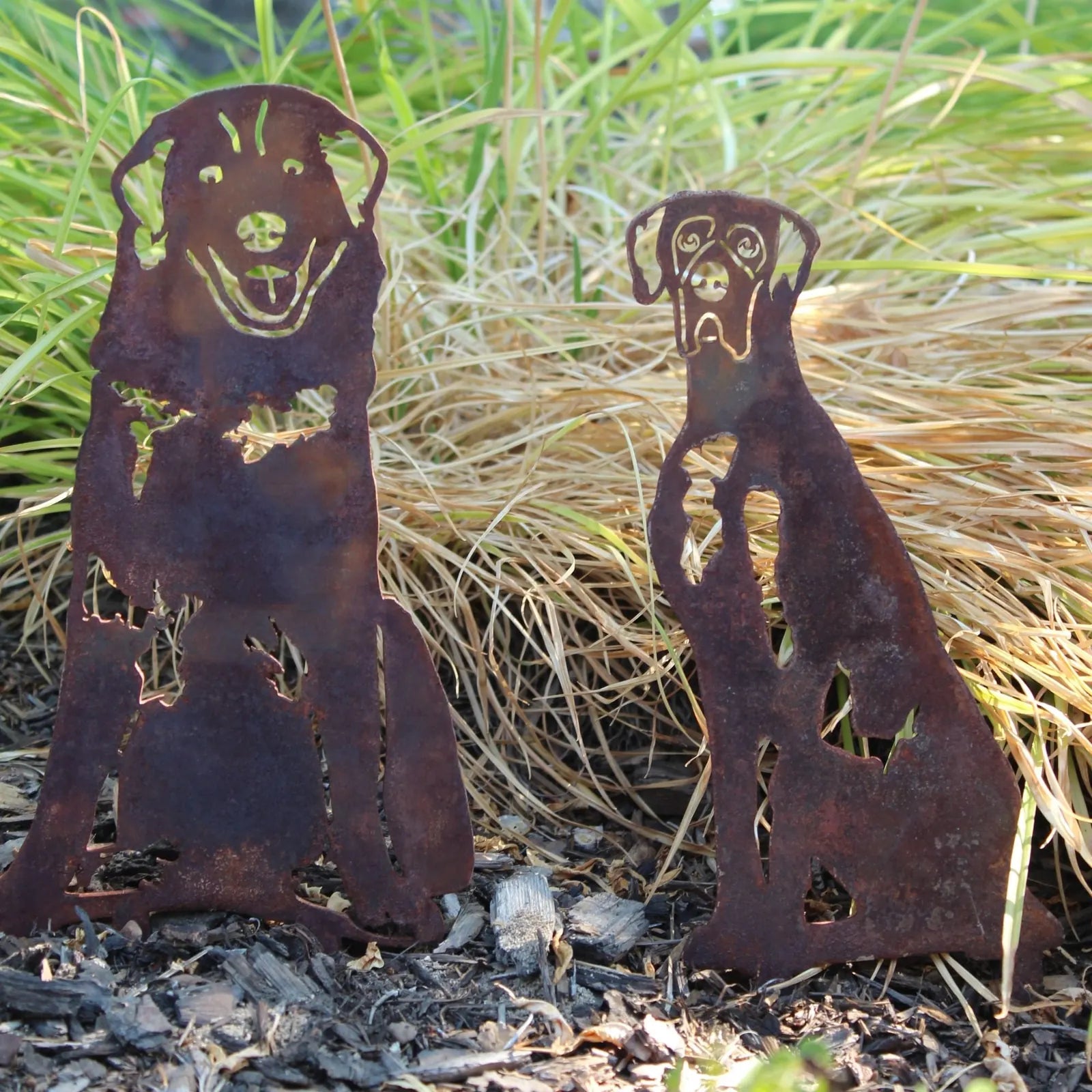 Rusty metal silhouette of a Great Pyrenees and smaller dog standing on mulch, surrounded by grass.