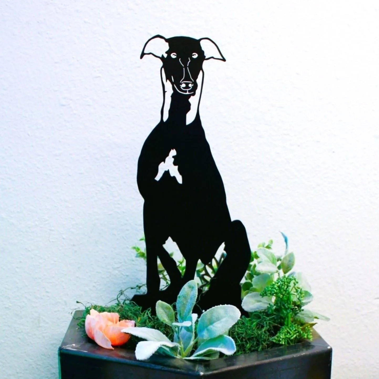 Black metal silhouette of a Greyhound dog sitting among green plants and flowers, perfect for outdoor decor.