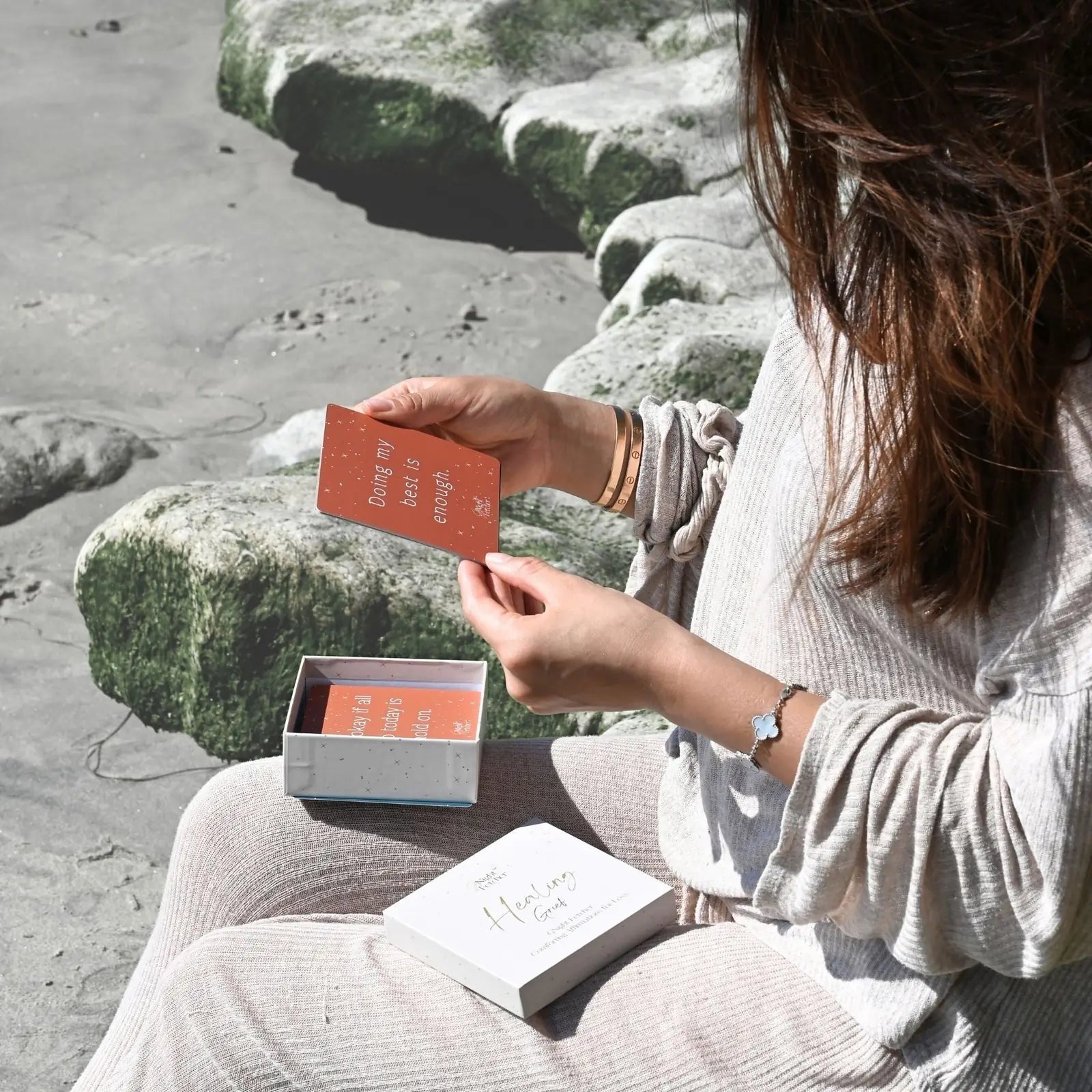 A person sitting on a beach holds a peach affirmation card, with a white box nearby containing more cards.