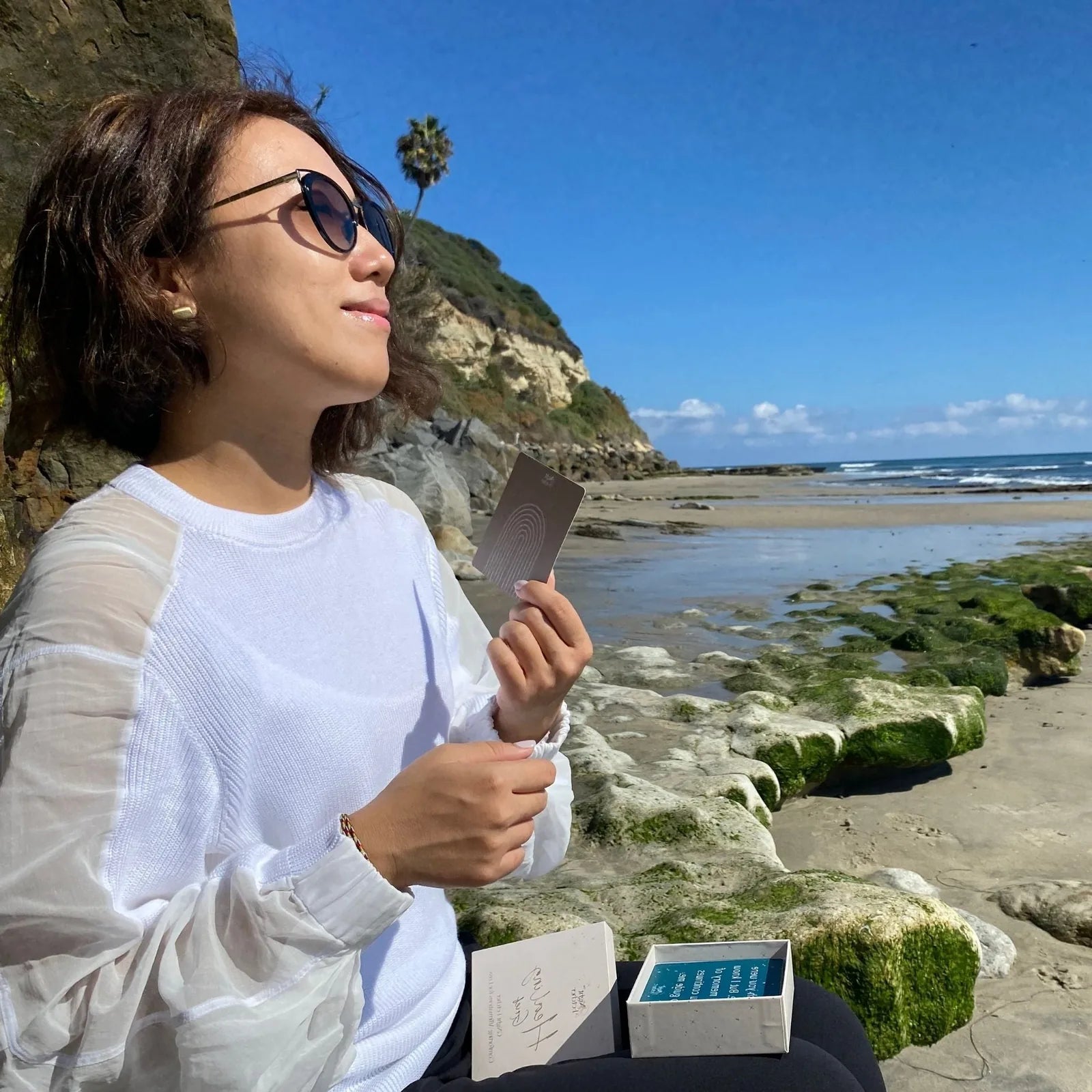 Woman wearing sunglasses holds a Grief Affirmation Card by the beach, with lush cliffs and ocean waves in the background.