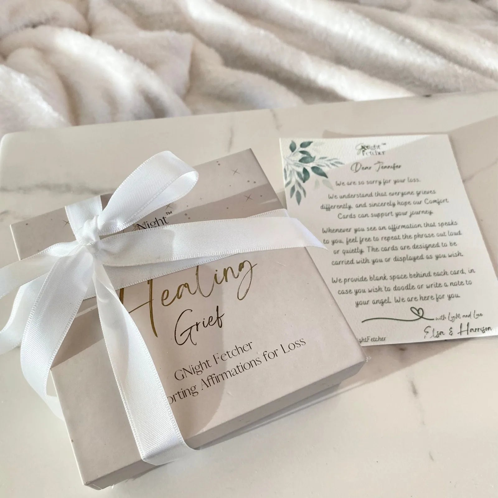 Grief Affirmation Cards in a cream box with a white ribbon, resting beside a sympathy note on a light surface.