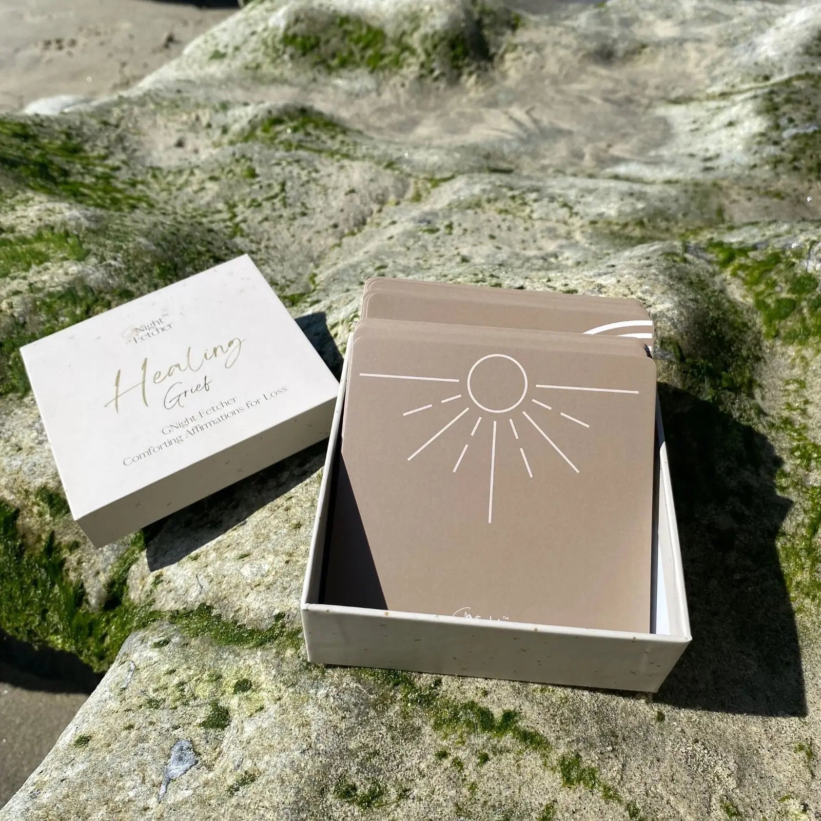 Grief affirmation cards in a box on a rocky surface, featuring a sun design on beige cards.