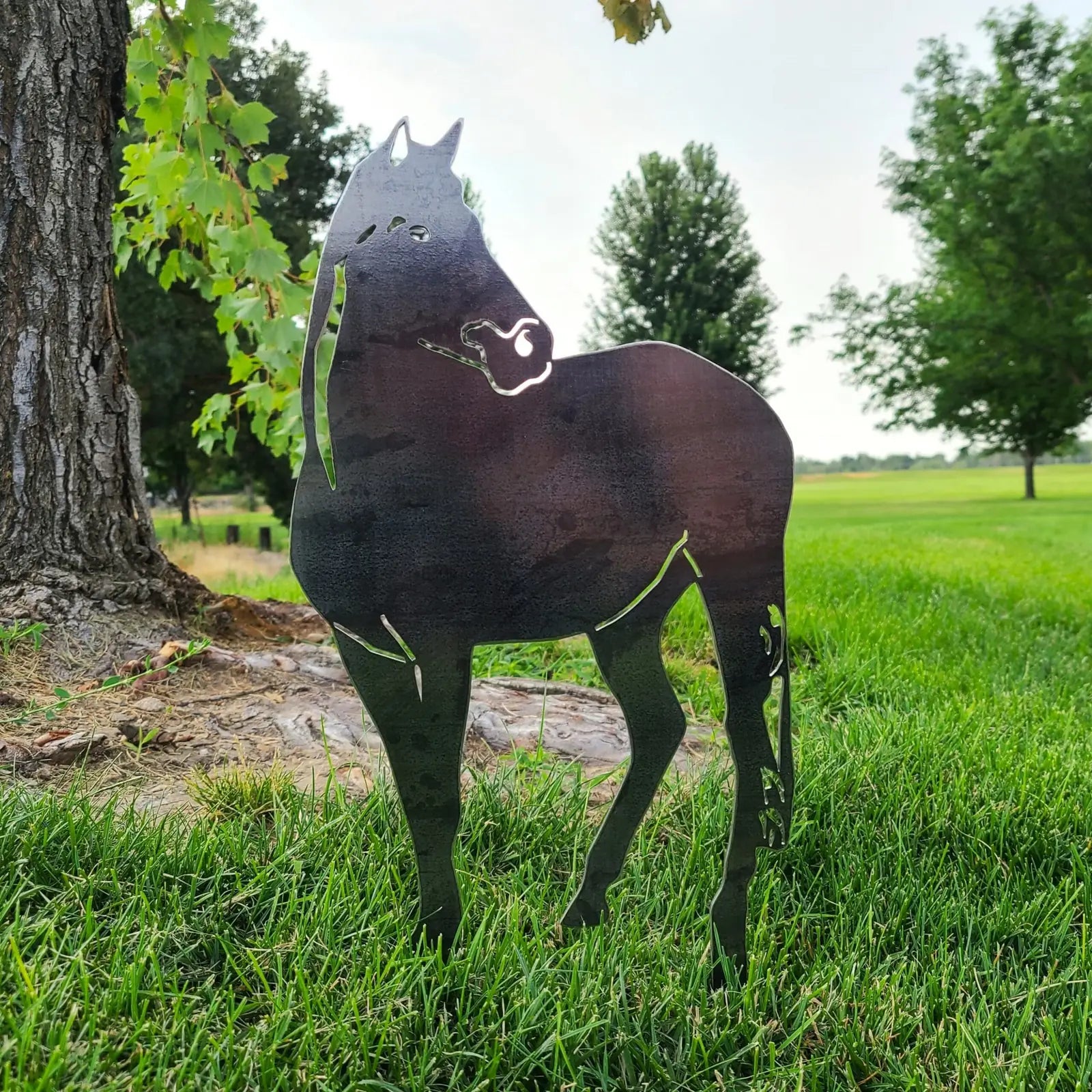 Black horse silhouette made from sturdy metal, standing on a grassy lawn with trees in the background.