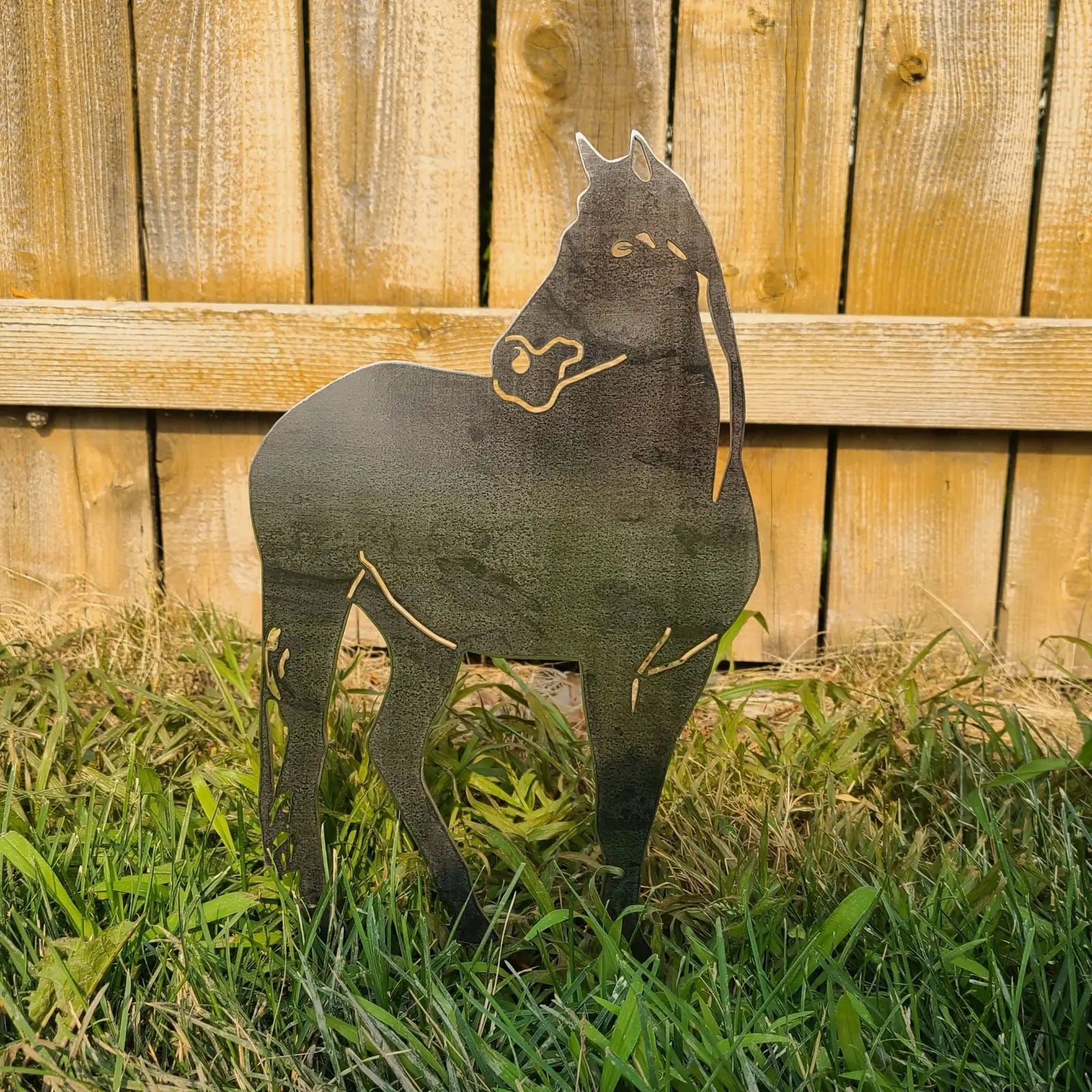 Corten steel horse silhouette stands in grass, showcasing intricate cutouts against a wooden fence backdrop.