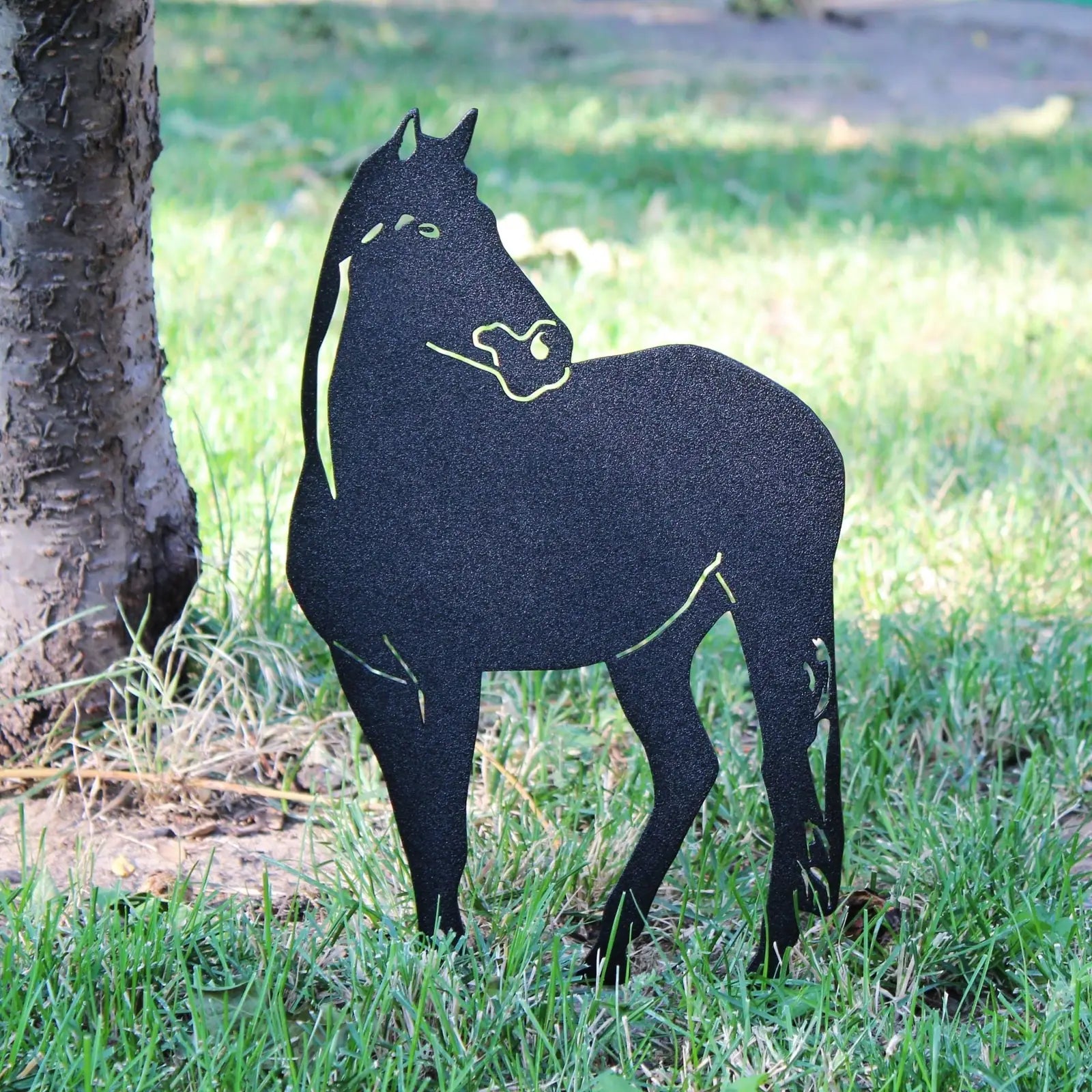 Black horse silhouette made from metal, standing on grassy ground near a tree, perfect for outdoor decor.