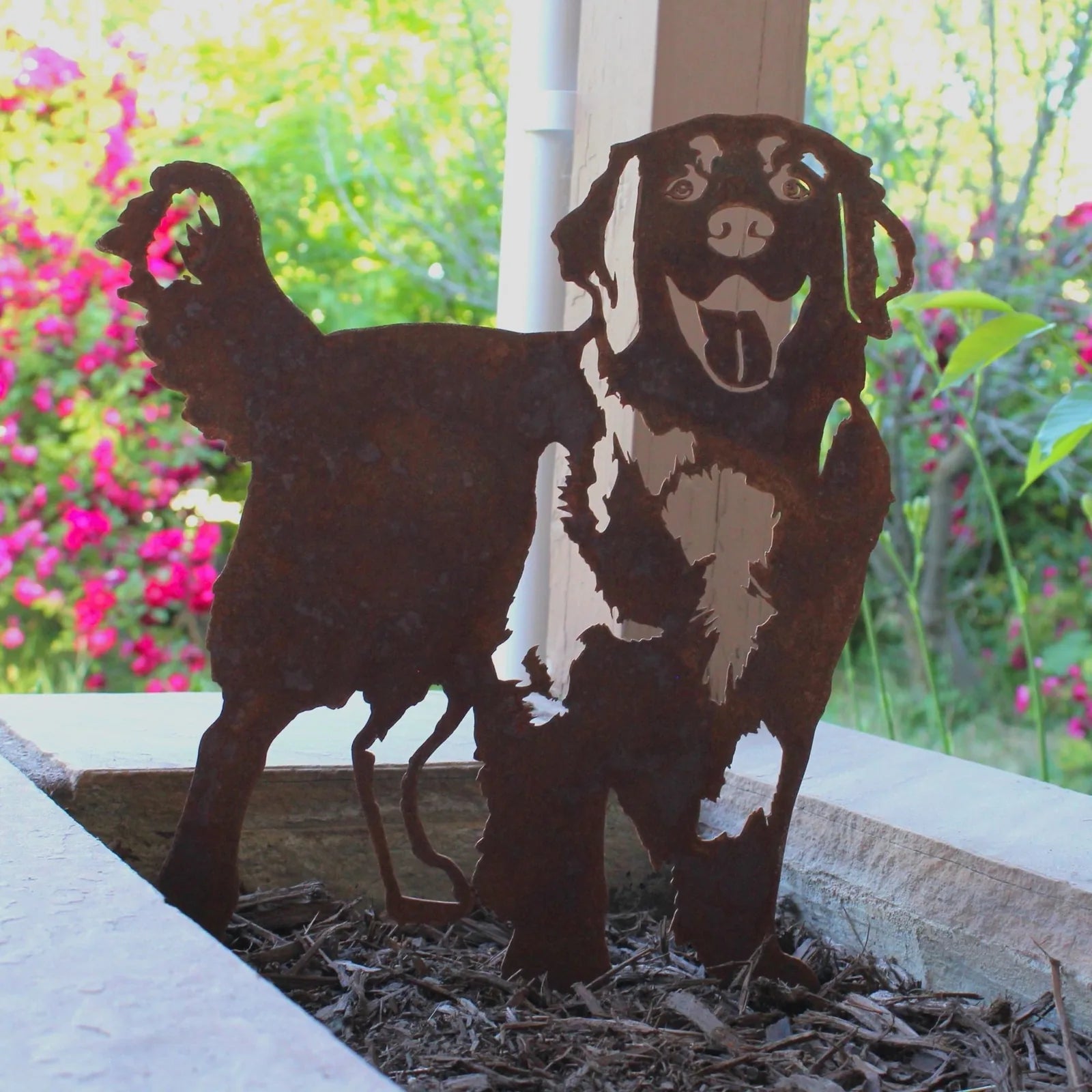 Hovawart dog silhouette made of rusty Corten steel, standing on mulch in a garden setting with colorful flowers.
