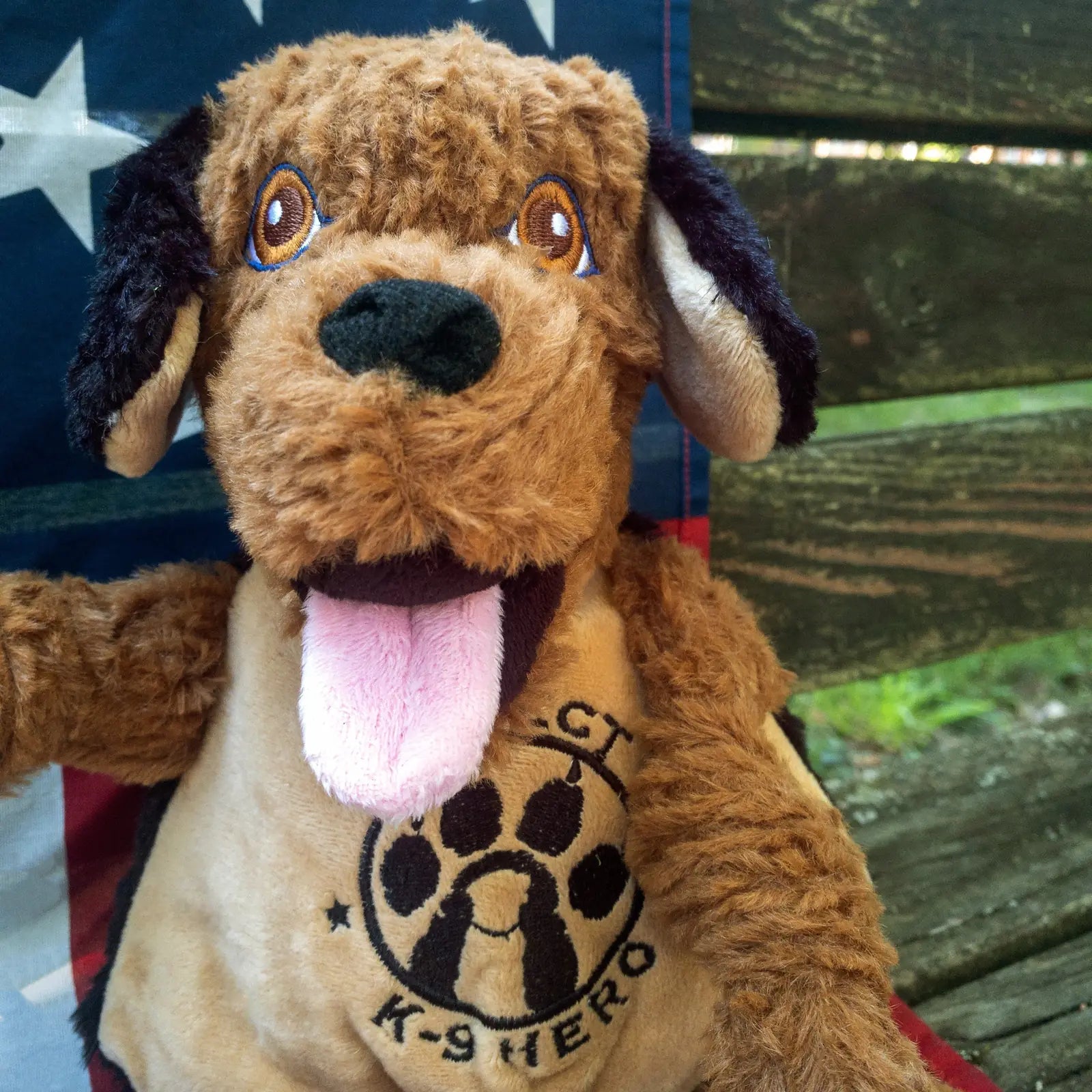 Plush brown dog toy with knotted limbs, stitched eyes, and a "Project K-9 Hero" logo, set against an American flag backdrop.