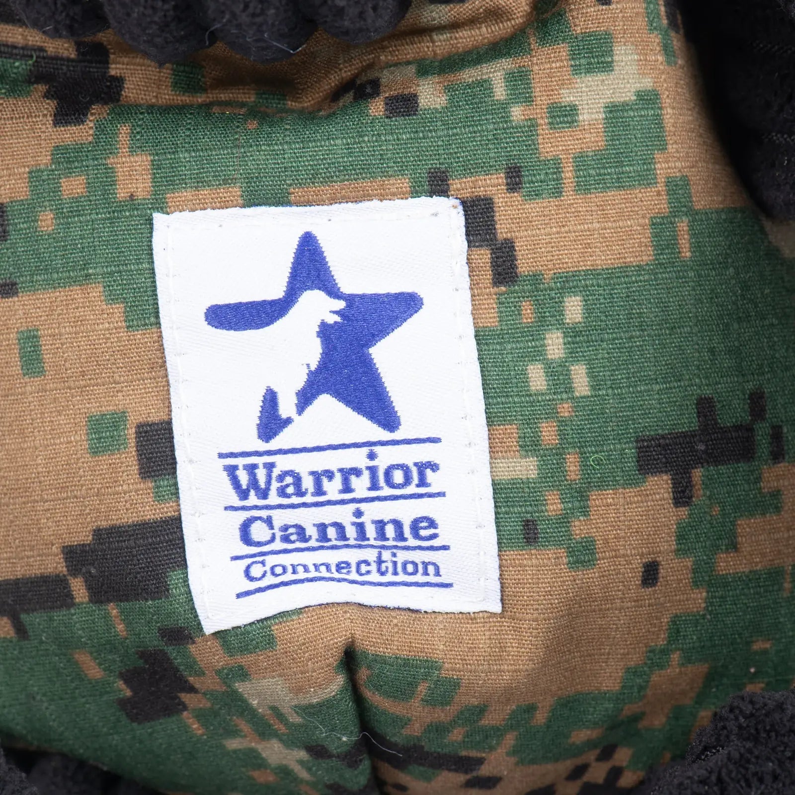 Warrior Canine Connection label on a plush dog toy featuring a camouflage pattern in greens and browns.