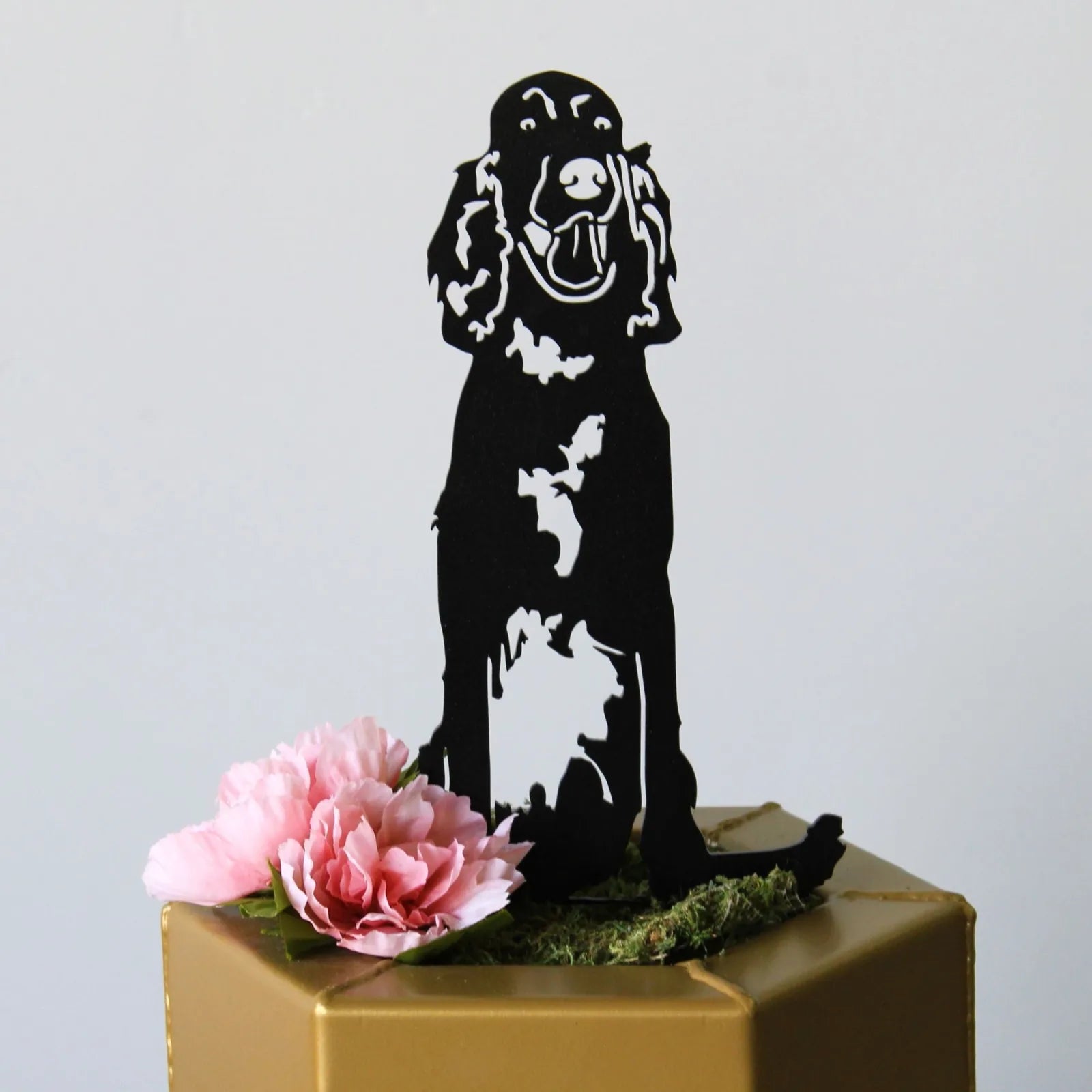 Black metal silhouette of an Irish Setter, sitting atop a decorated base with pink flowers and moss.