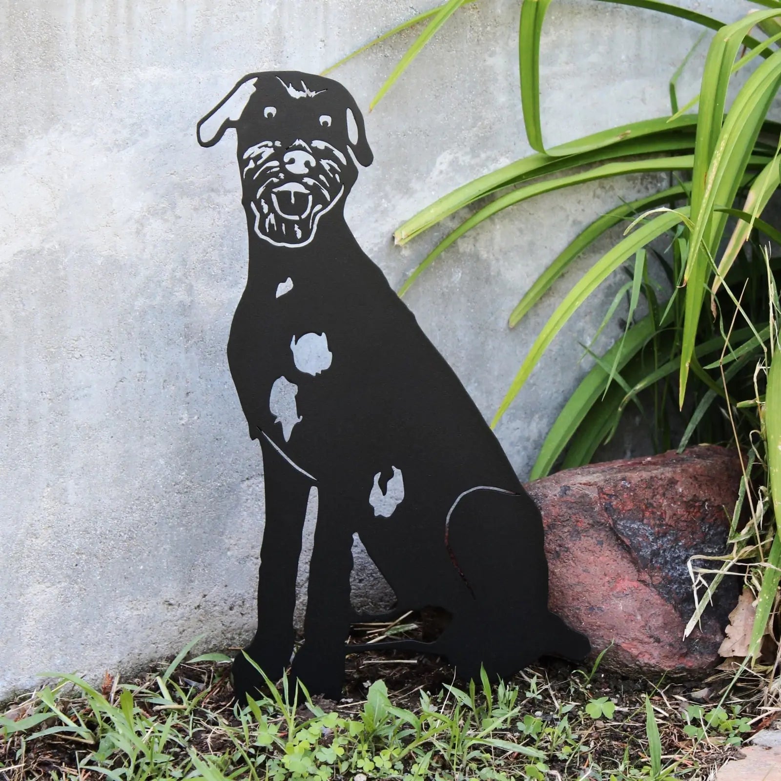 Black metal silhouette of an Irish Wolfhound sitting among green foliage and a stone, showcasing detailed craftsmanship.