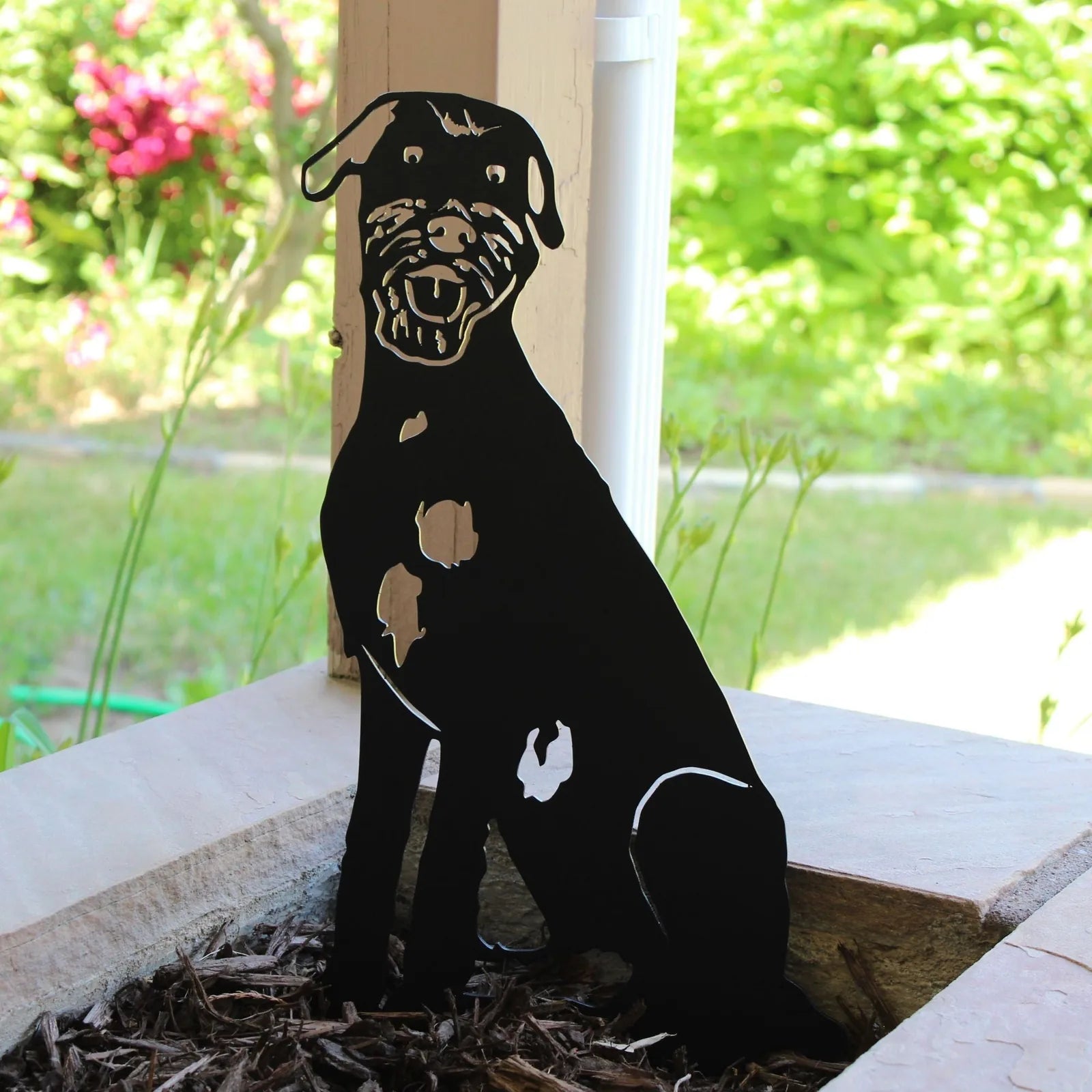 Black metal silhouette of an Irish Wolfhound resting in garden mulch, with greenery in the background.