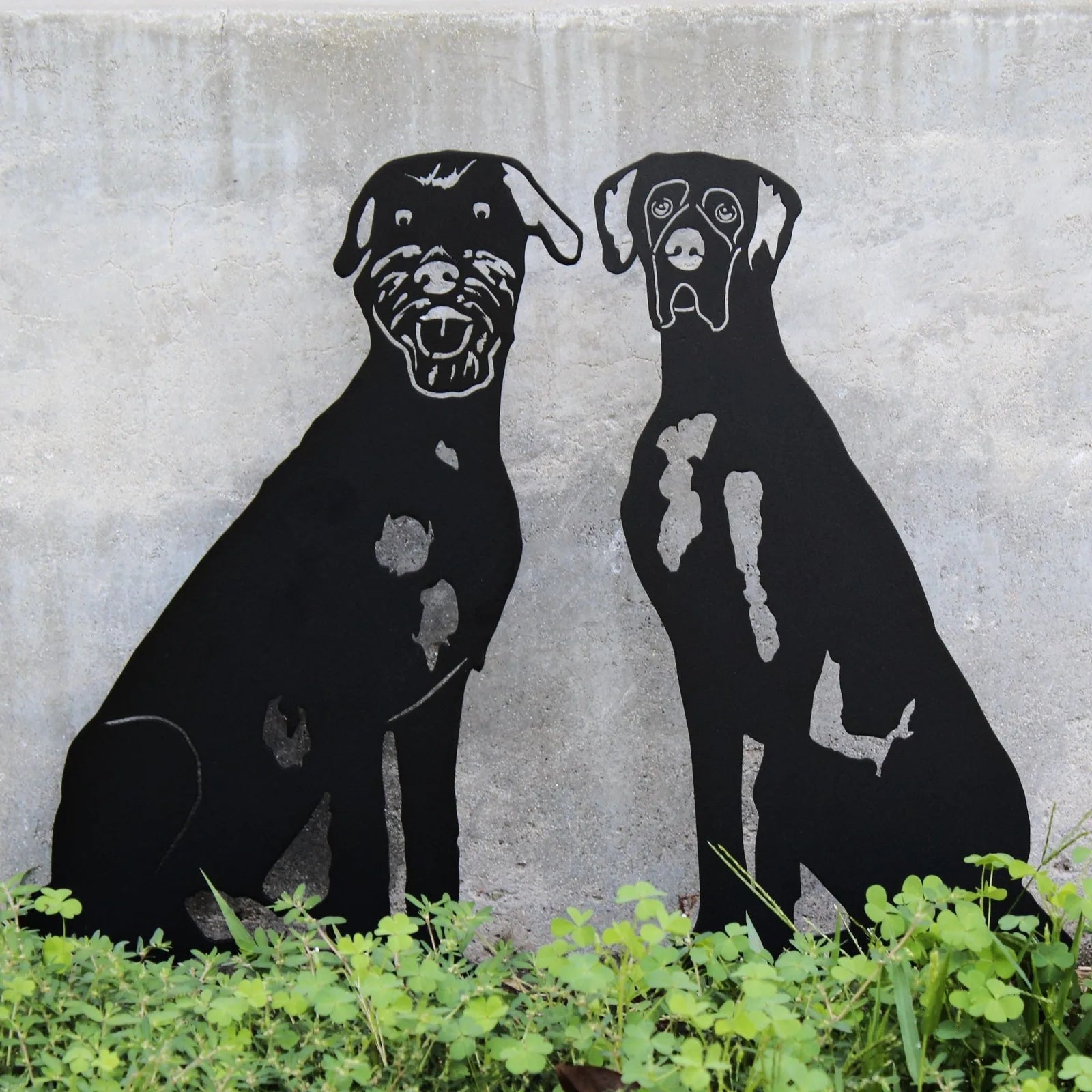 Black metal silhouette of two dogs sitting against a concrete wall, surrounded by green plants, showcasing outdoor decor.