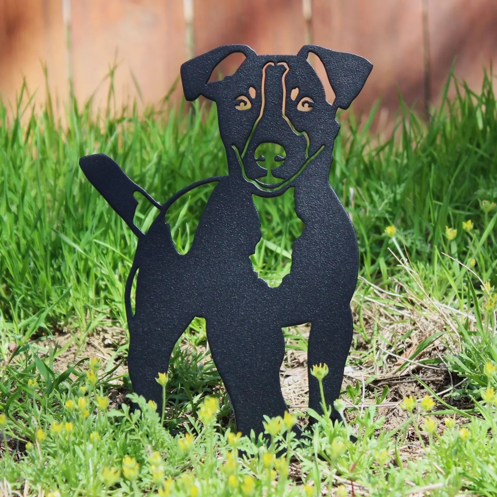 Black Jack Russell Terrier metal silhouette art piece, set among green grass and wildflowers, showcasing outdoor decor style.