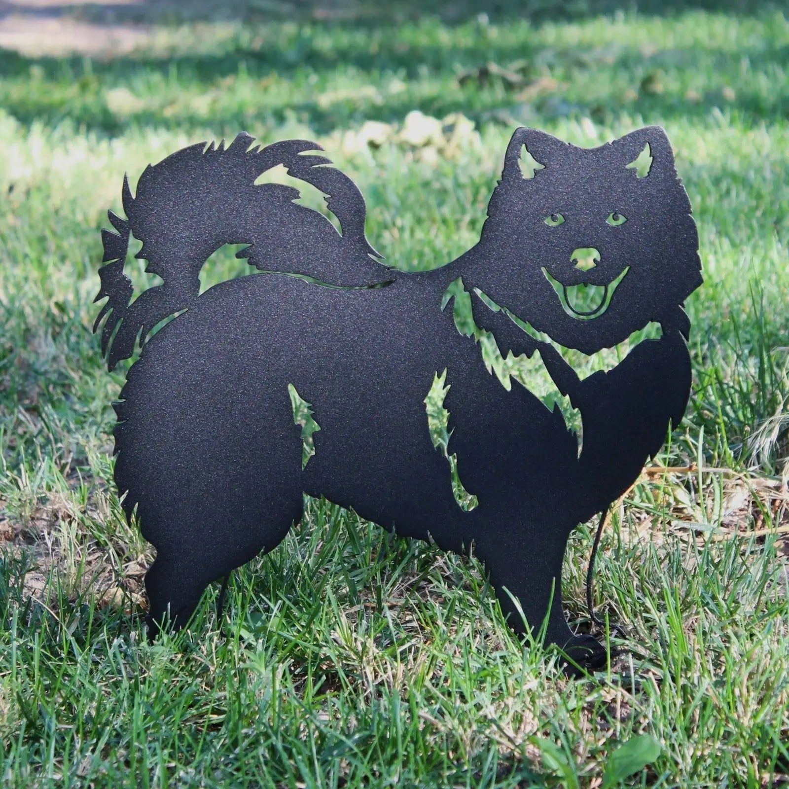 Black metal silhouette of a Japanese Spitz dog standing on green grass, showcasing detailed features and a playful design.