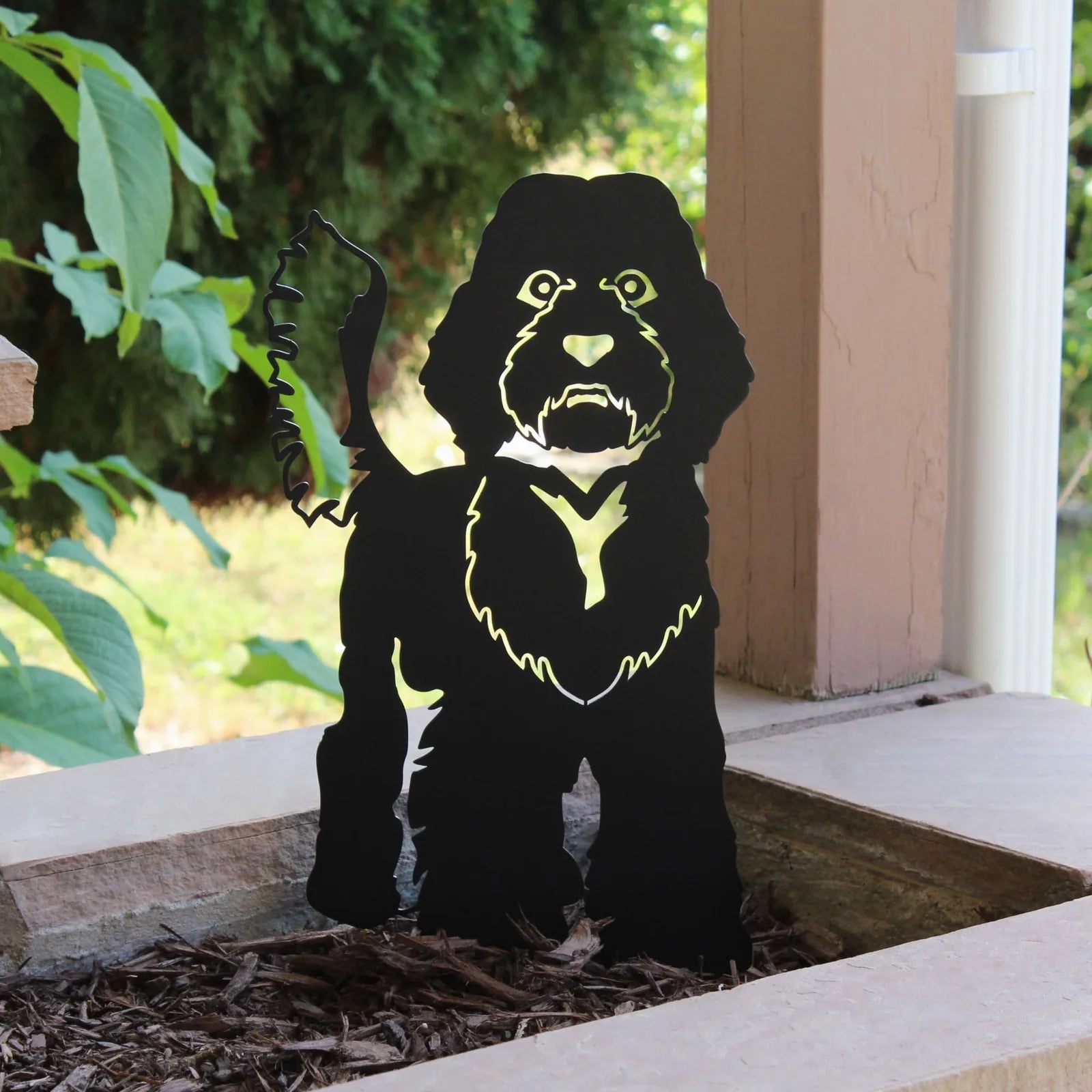 Handmade black metal silhouette of a Labradoodle installed in garden mulch, surrounded by greenery.