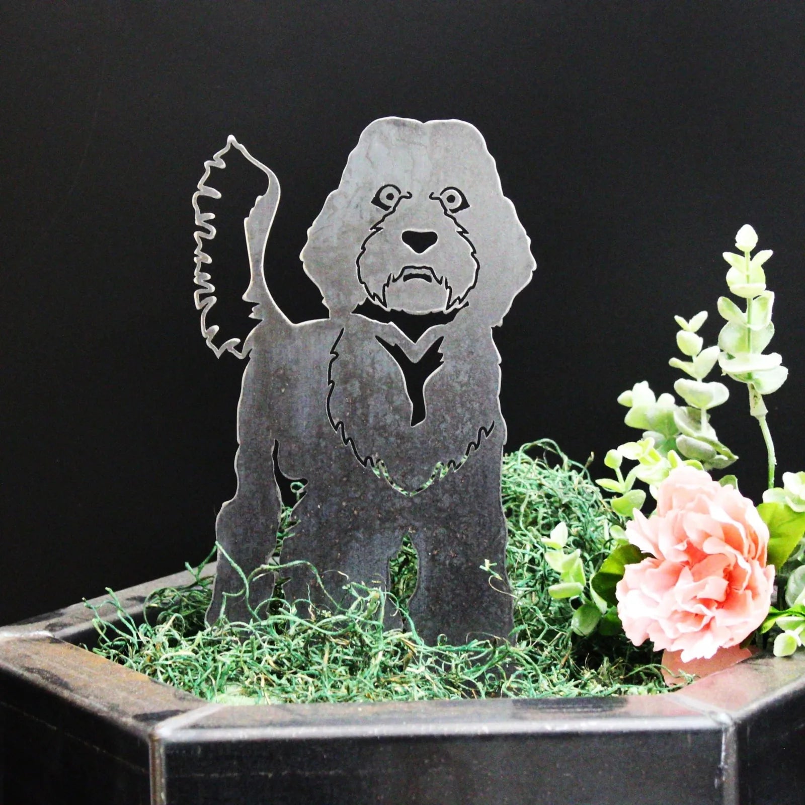 Labradoodle metal silhouette in black, set among green moss and flowers, showcasing a rustic outdoor decoration.
