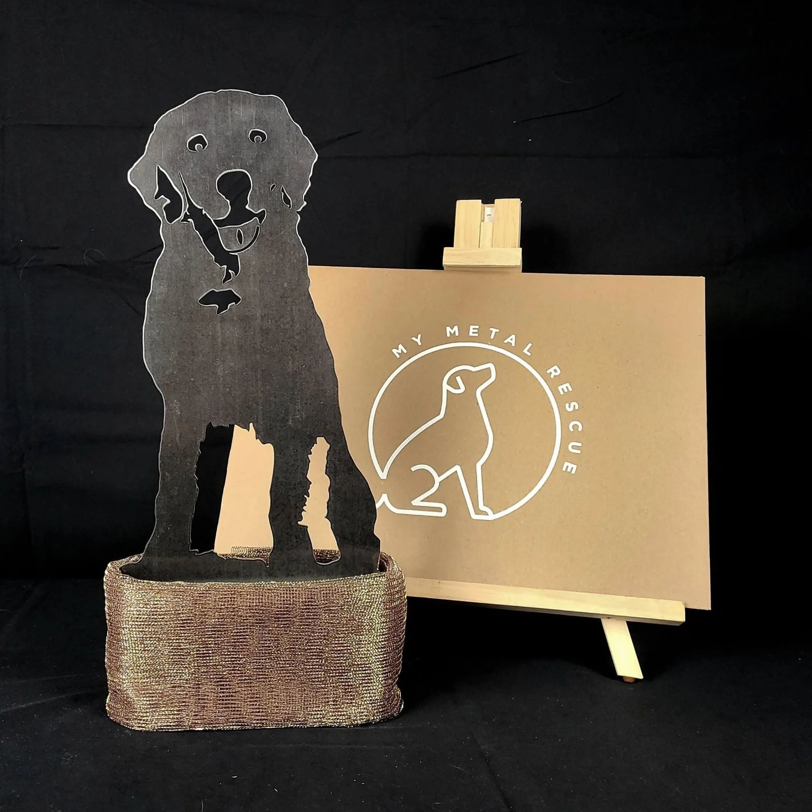 Handcrafted black metal silhouette of a Labrador Retriever resting on a textured, metallic base, with branding in the back...