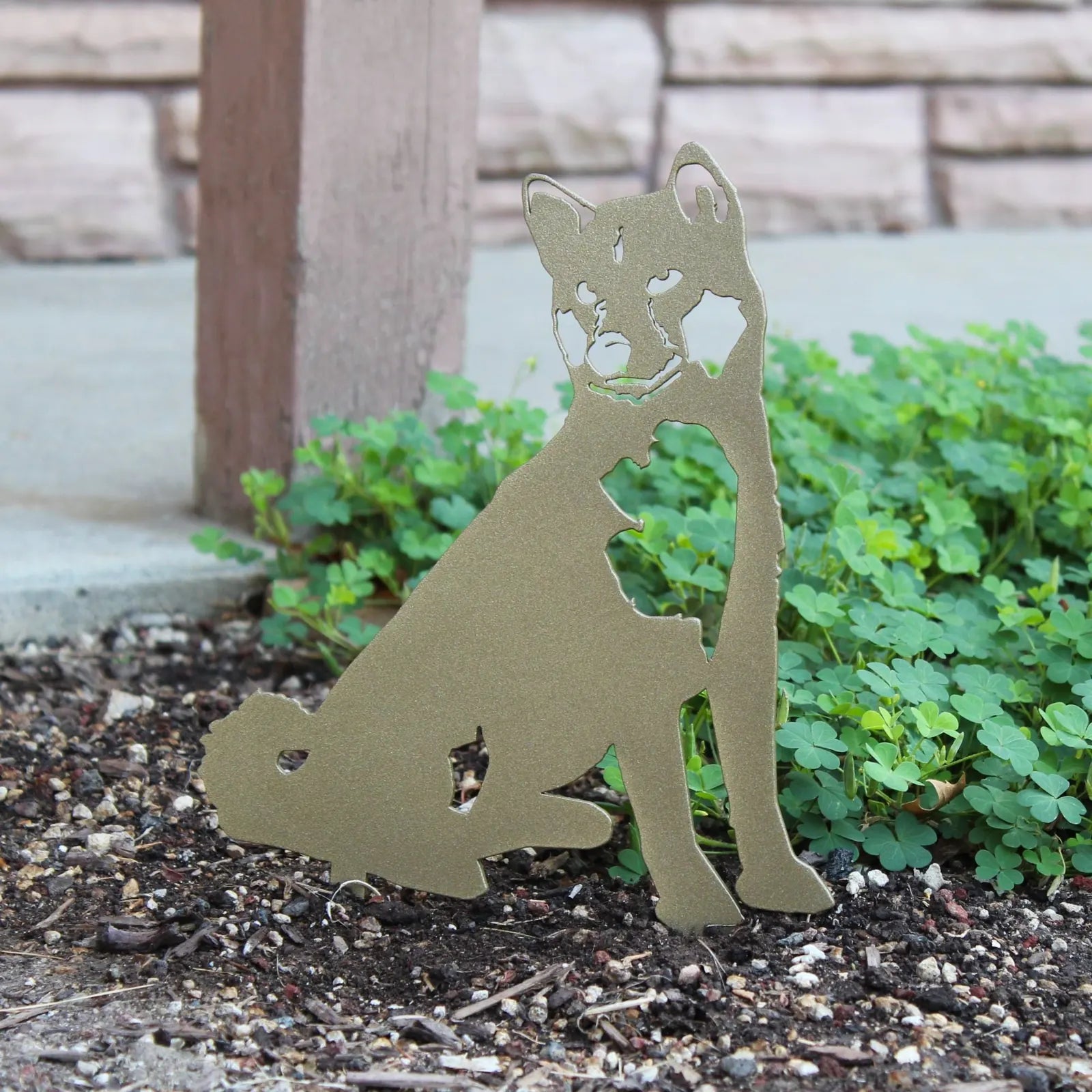 Handmade metallic gold Shiba Inu silhouette in weathering steel, displayed in a garden setting among green plants.