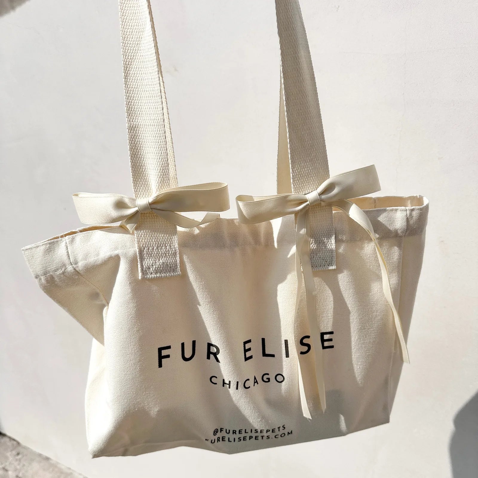 Cream canvas tote bag with wide straps and bow accents, featuring black text branding for "FUR ELISE CHICAGO."