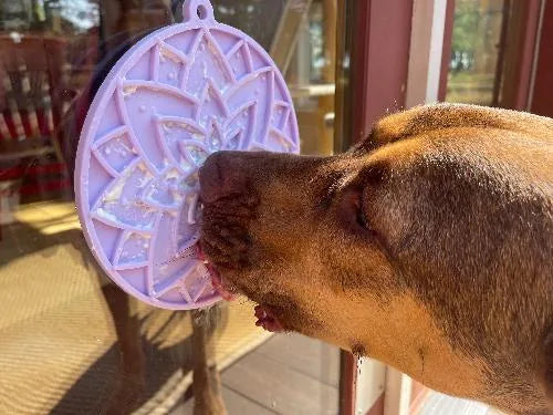 Dog enjoying a pastel purple lotus design lick mat attached to a glass surface, licking off soft treats.