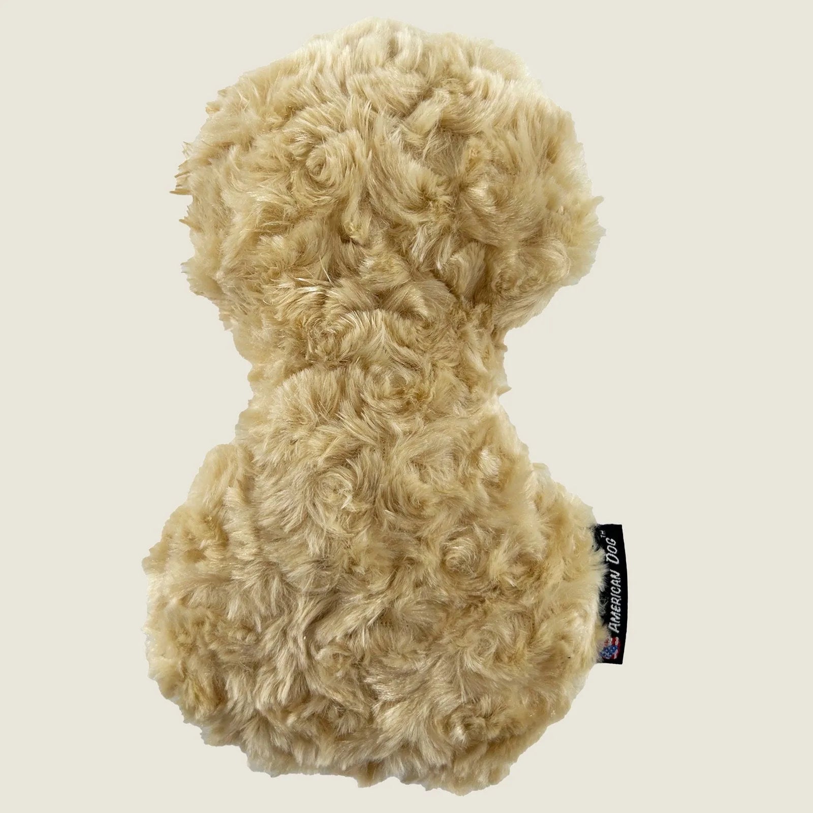 Soft, plush peanut-shaped dog toy in light tan with textured fur and an American Dog label, ideal for gentle play.