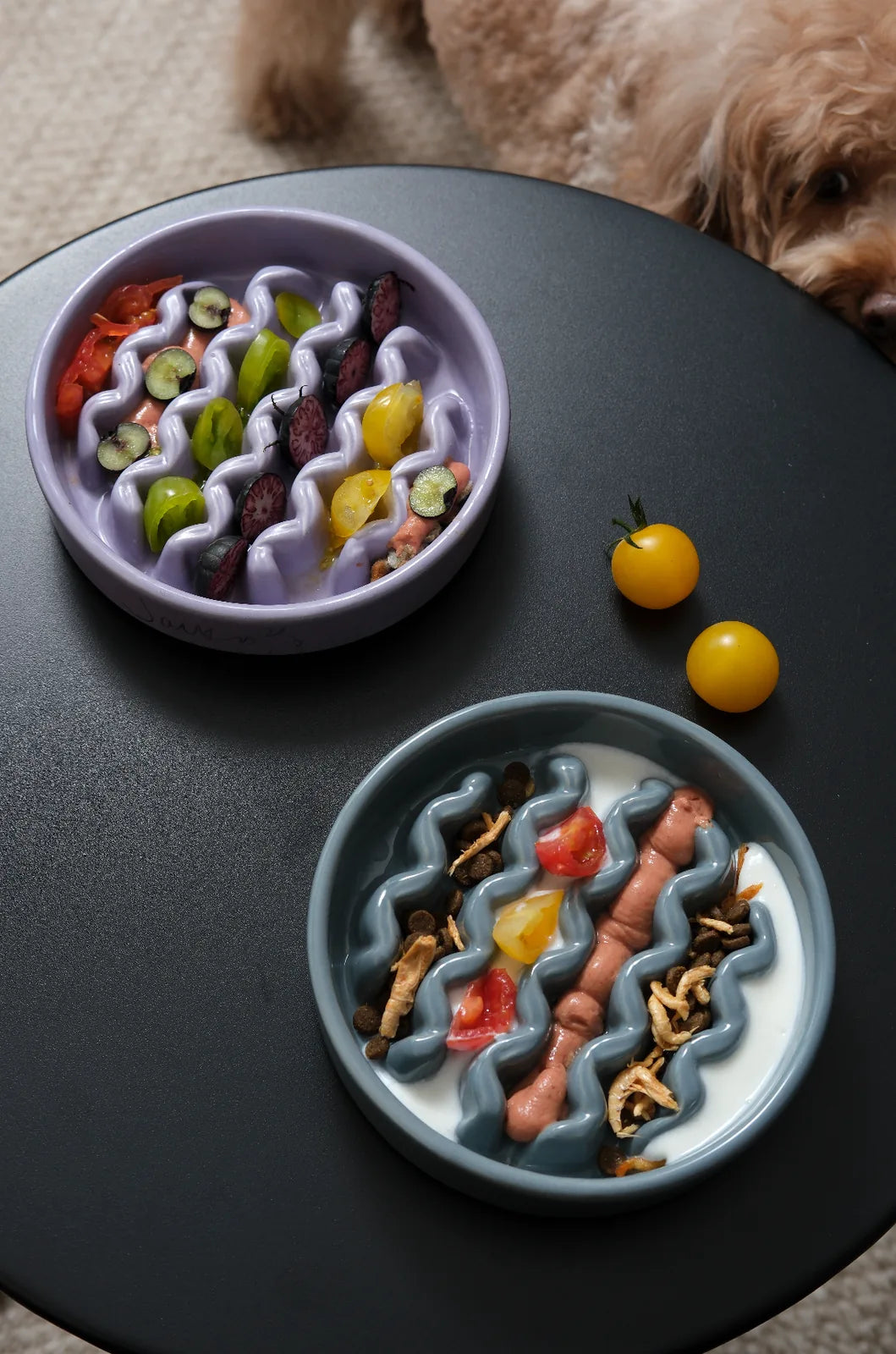 Purple and gray slow feeder dog bowls filled with various foods, displayed on a dark surface with yellow tomatoes nearby.