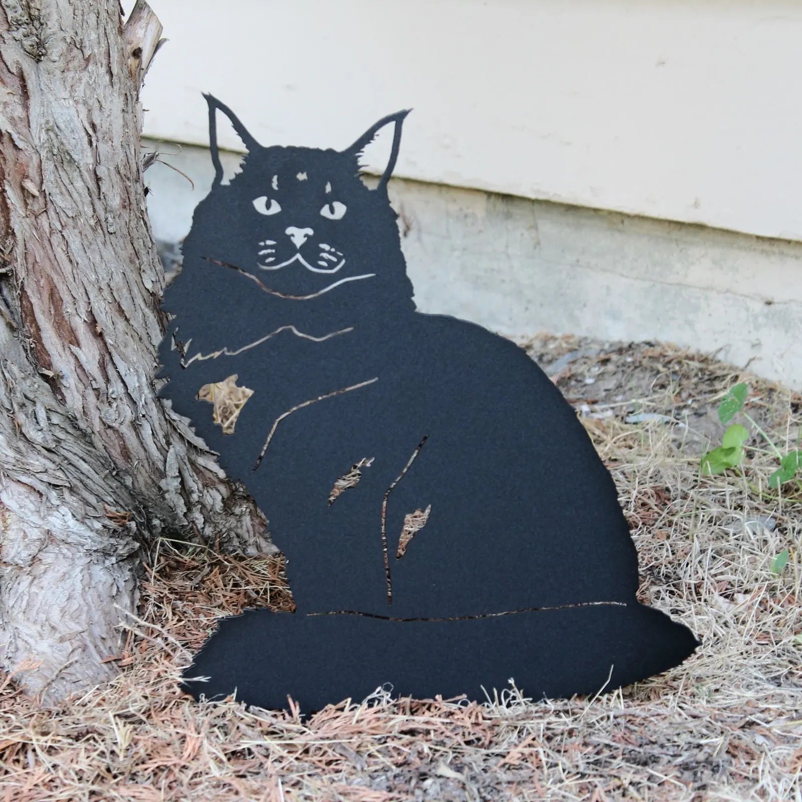 Black metal silhouette of a Maine Coon cat resting by a tree, showcasing intricate detailing in a rustic garden setting.
