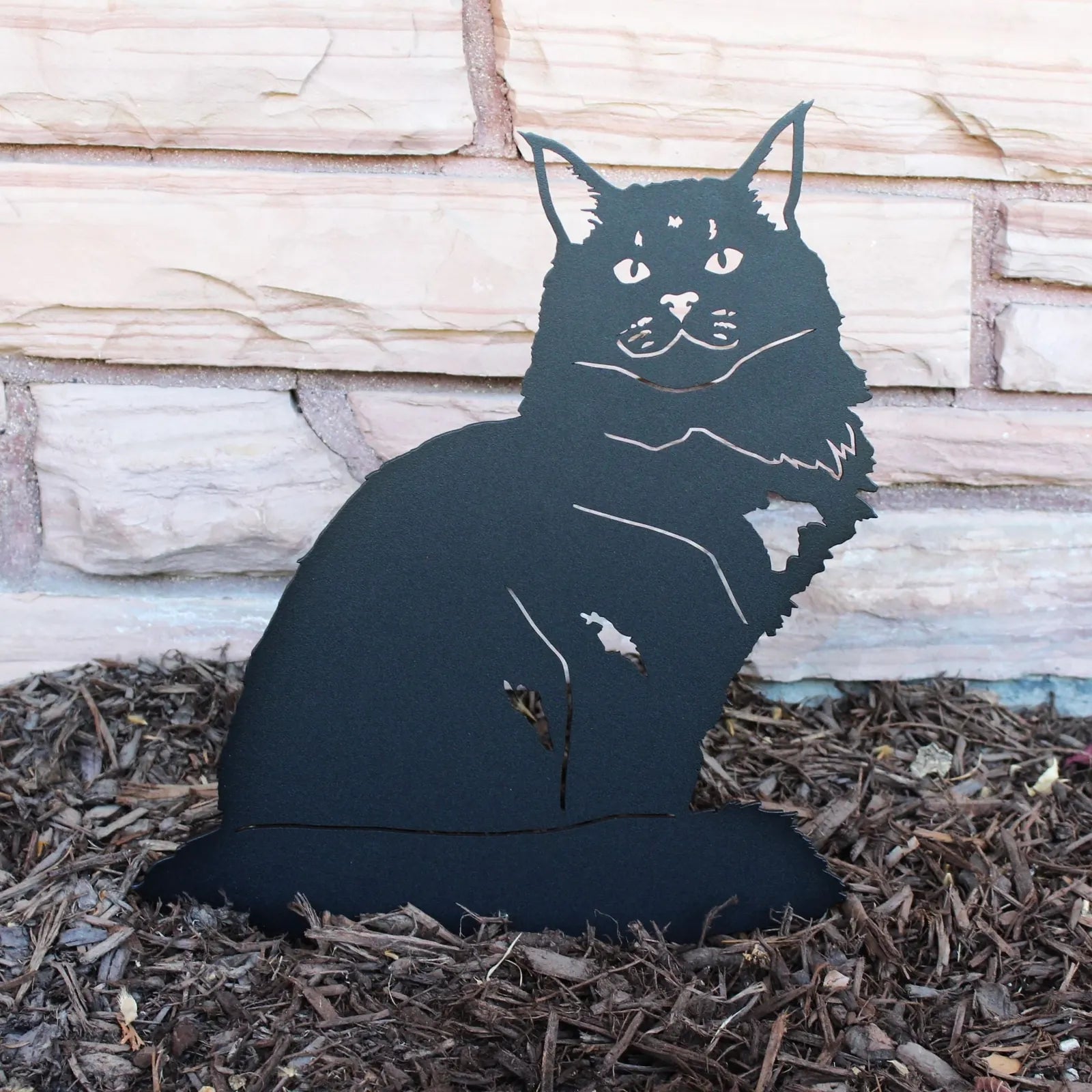 Black Maine Coon metal silhouette made from sturdy steel, set against mulch and a stone wall.