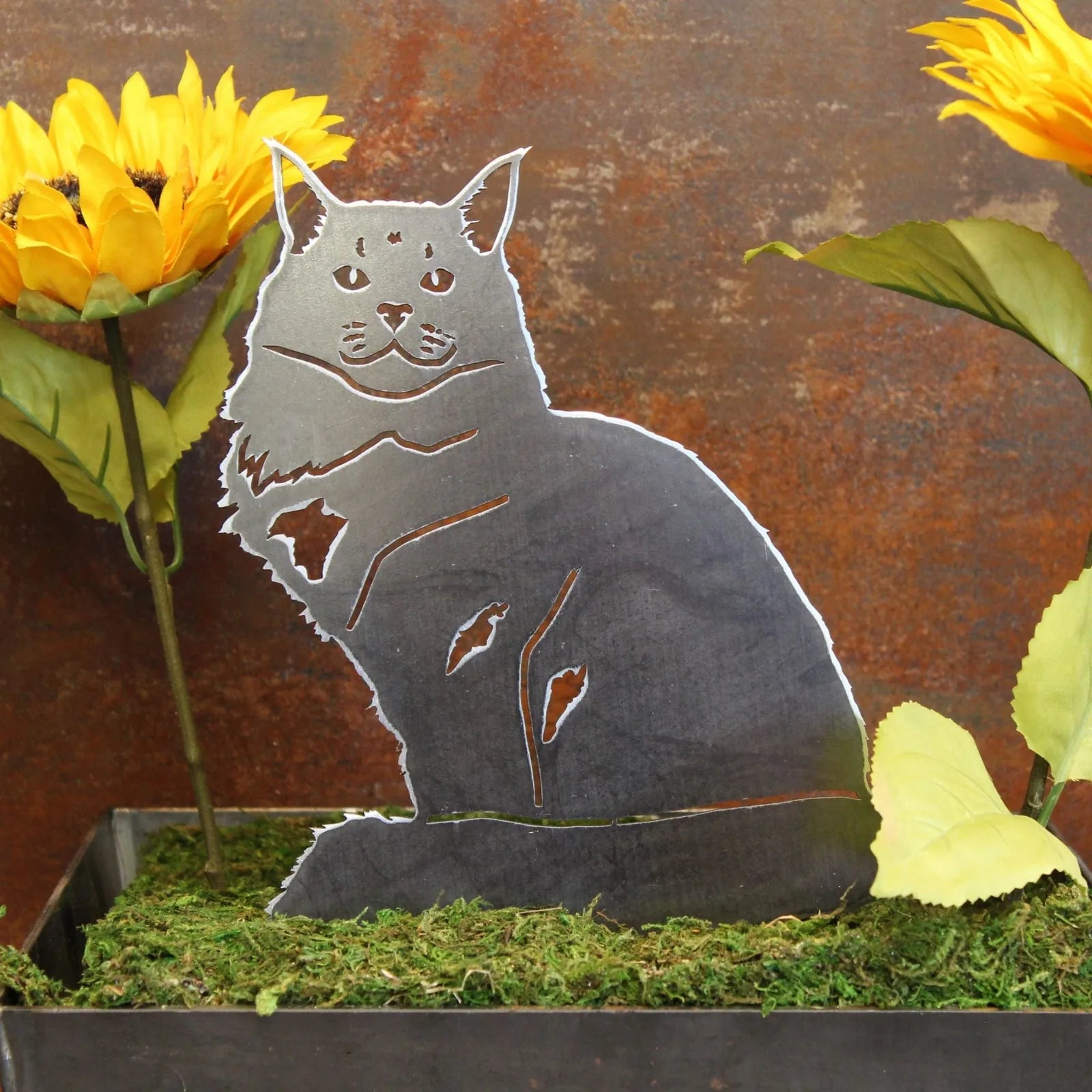 Maine Coon metal silhouette in matte black, surrounded by sunflowers and greenery in a rustic outdoor setting.