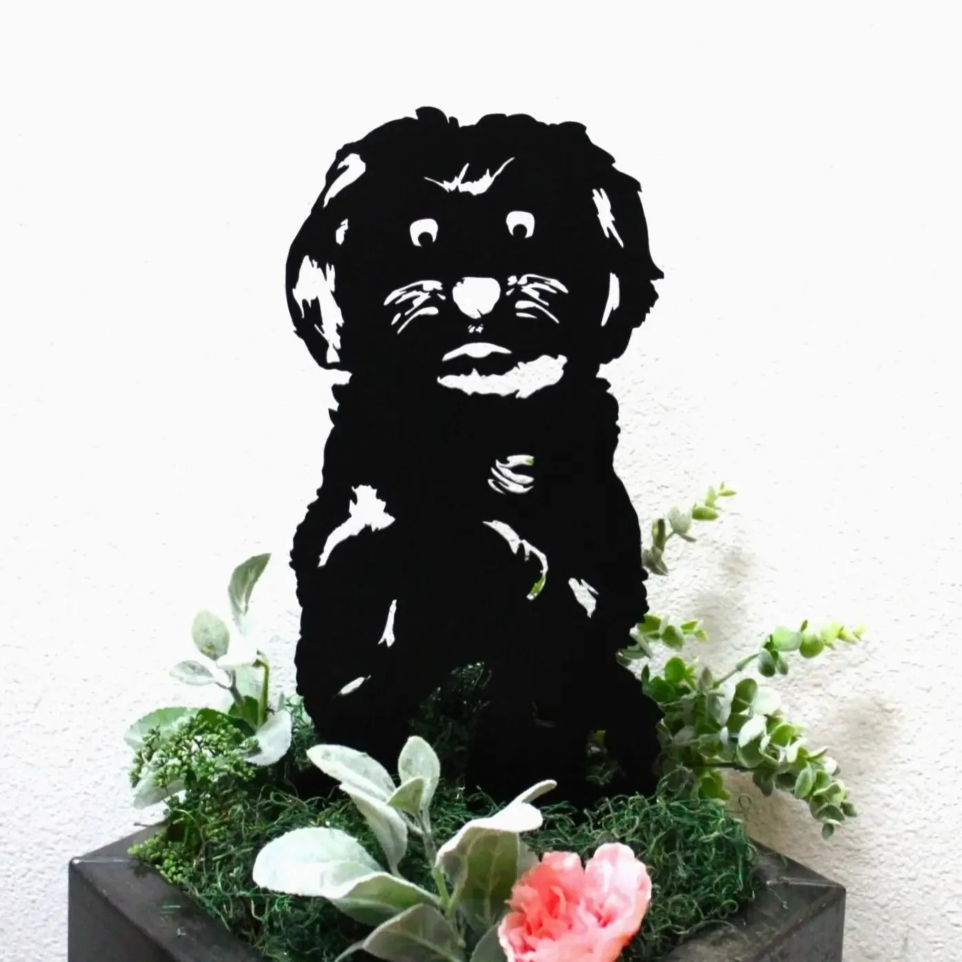 Black Maltese metal silhouette art piece surrounded by greenery and blooms, perfect for outdoor dog decor.