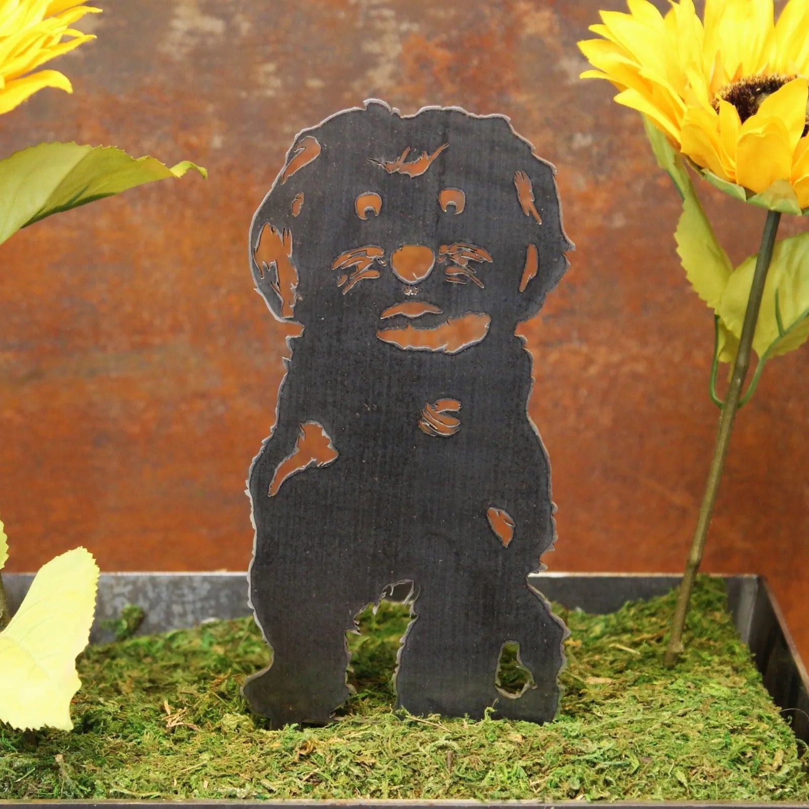 Black Maltese metal silhouette on moss, surrounded by yellow sunflowers against a rustic background.