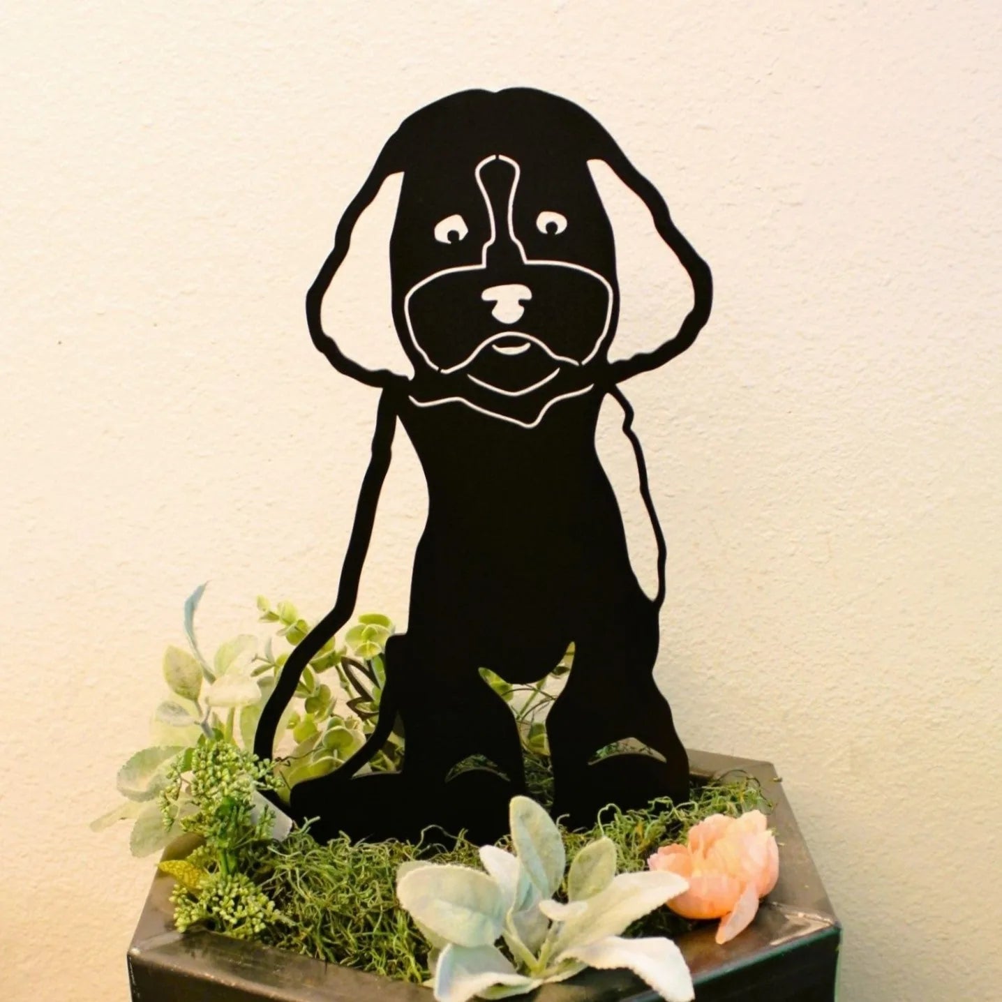 Black Maltipoo metal silhouette art placed among greenery and flowers, perfect for outdoor dog-themed decor.
