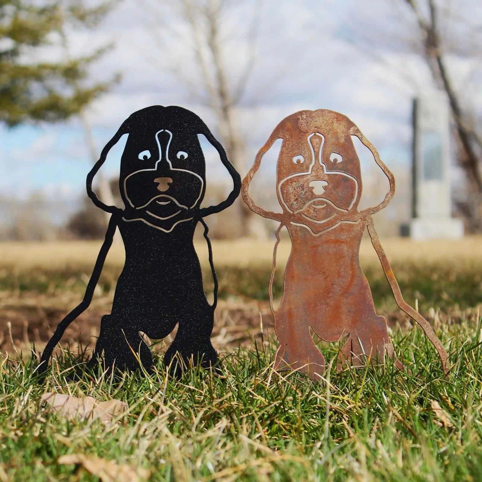 Two dog silhouette sculptures, one black and one rust-colored, standing in grass, showcasing Maltipoo design in metal art.